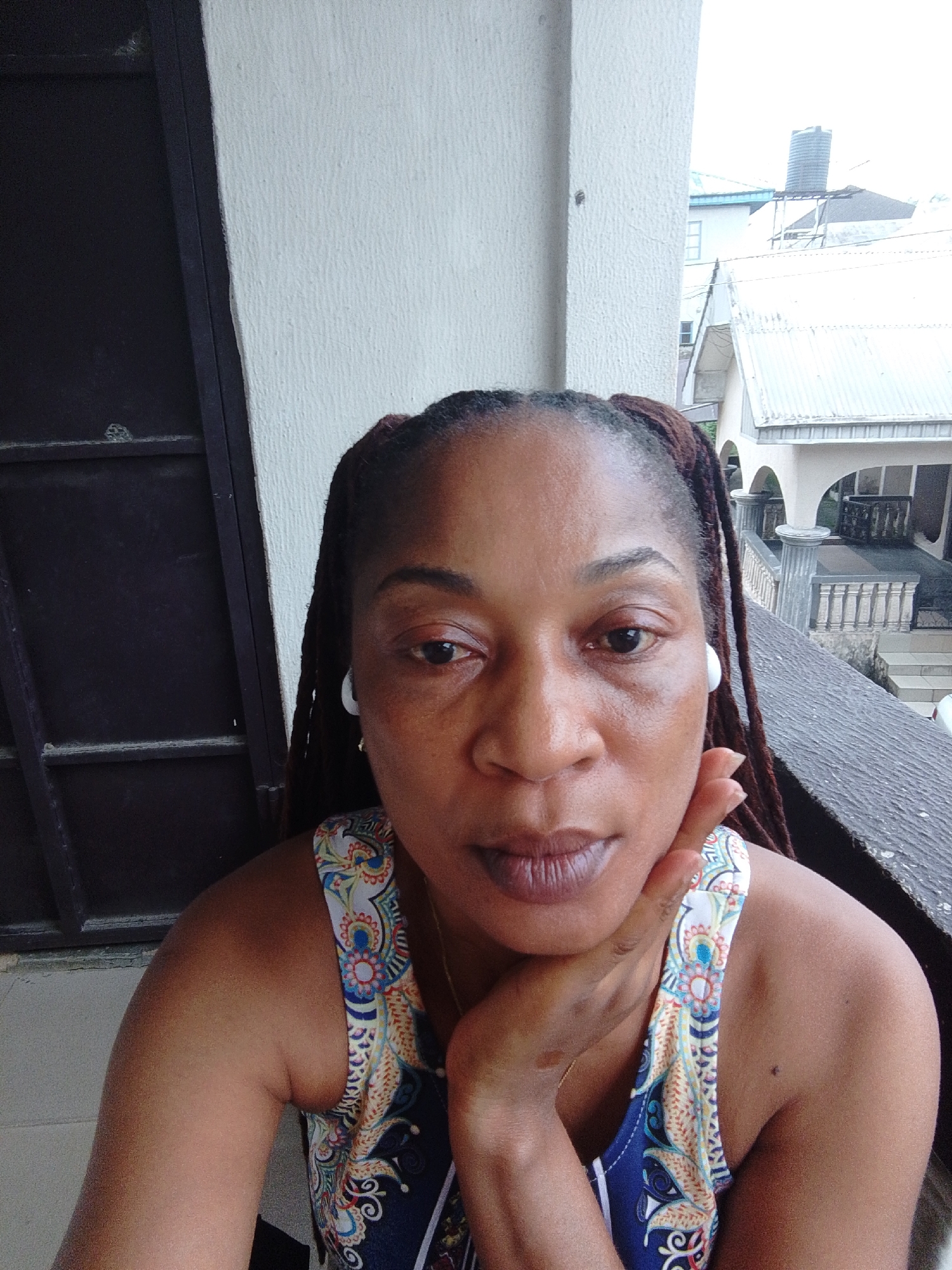 A medium shot image shows a Black woman with braided hair posing on a balcony in Calabar, Nigeria. The woman is wearing a blue and white patterned tank top and white earbuds. Her right hand is resting on her chin, and she is looking directly at the camera. The background shows a white textured wall and a railing of a balcony. Beyond the railing, there are residential buildings and a water tower. The lighting suggests it is daytime, possibly overcast.