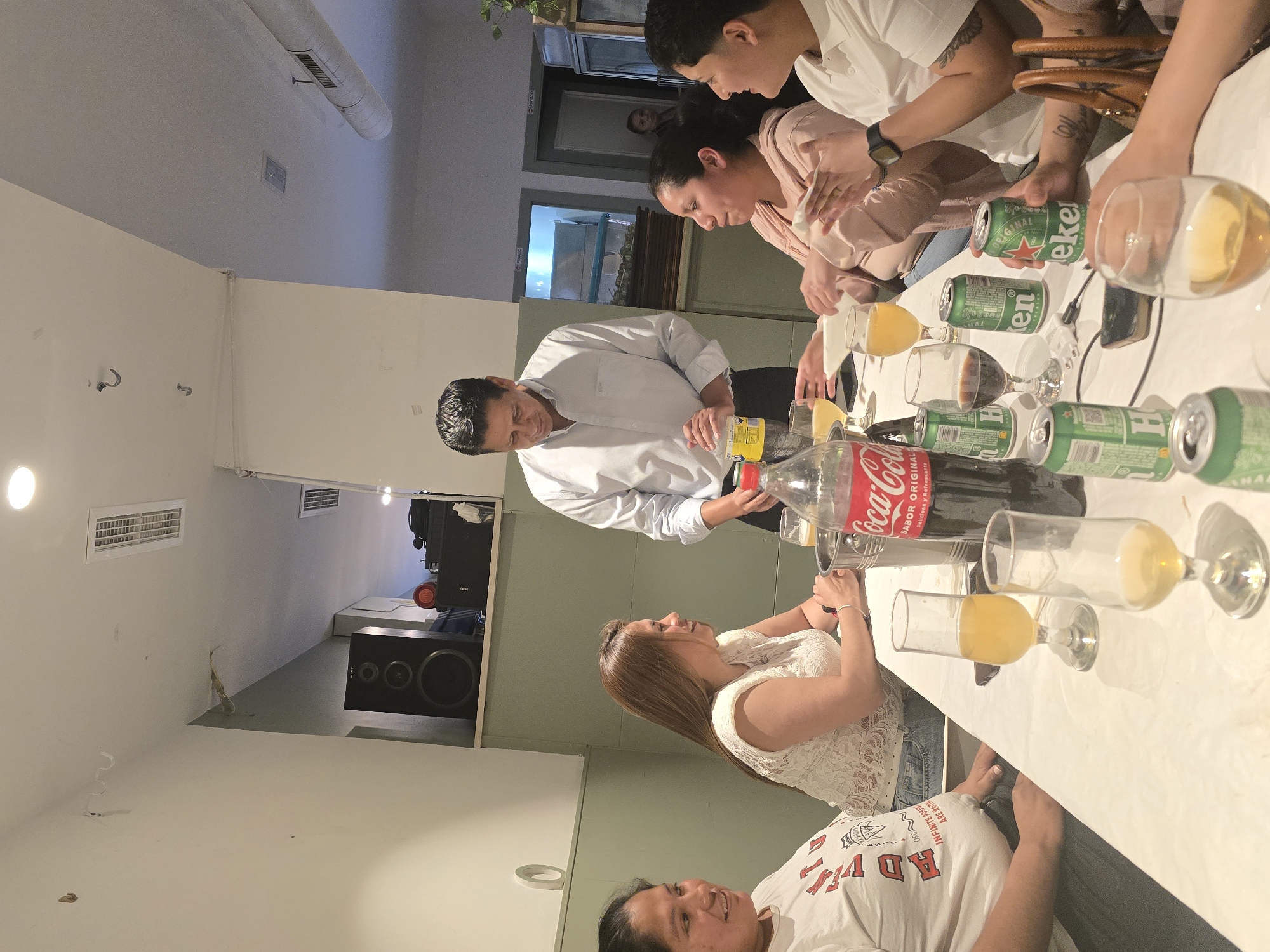 The image shows a gathering of people around a table, appearing to be socializing and enjoying drinks. There are several cans and bottles of beverages, including Heineken beer and Coca-Cola, along with glasses filled with liquids. The scene is indoors, potentially in a home or event space. The presence of a speaker and subwoofer system suggests a recreational or party-like atmosphere. The people in the image seem to be engaged in conversation and interaction. Location cannot be determined from the image.