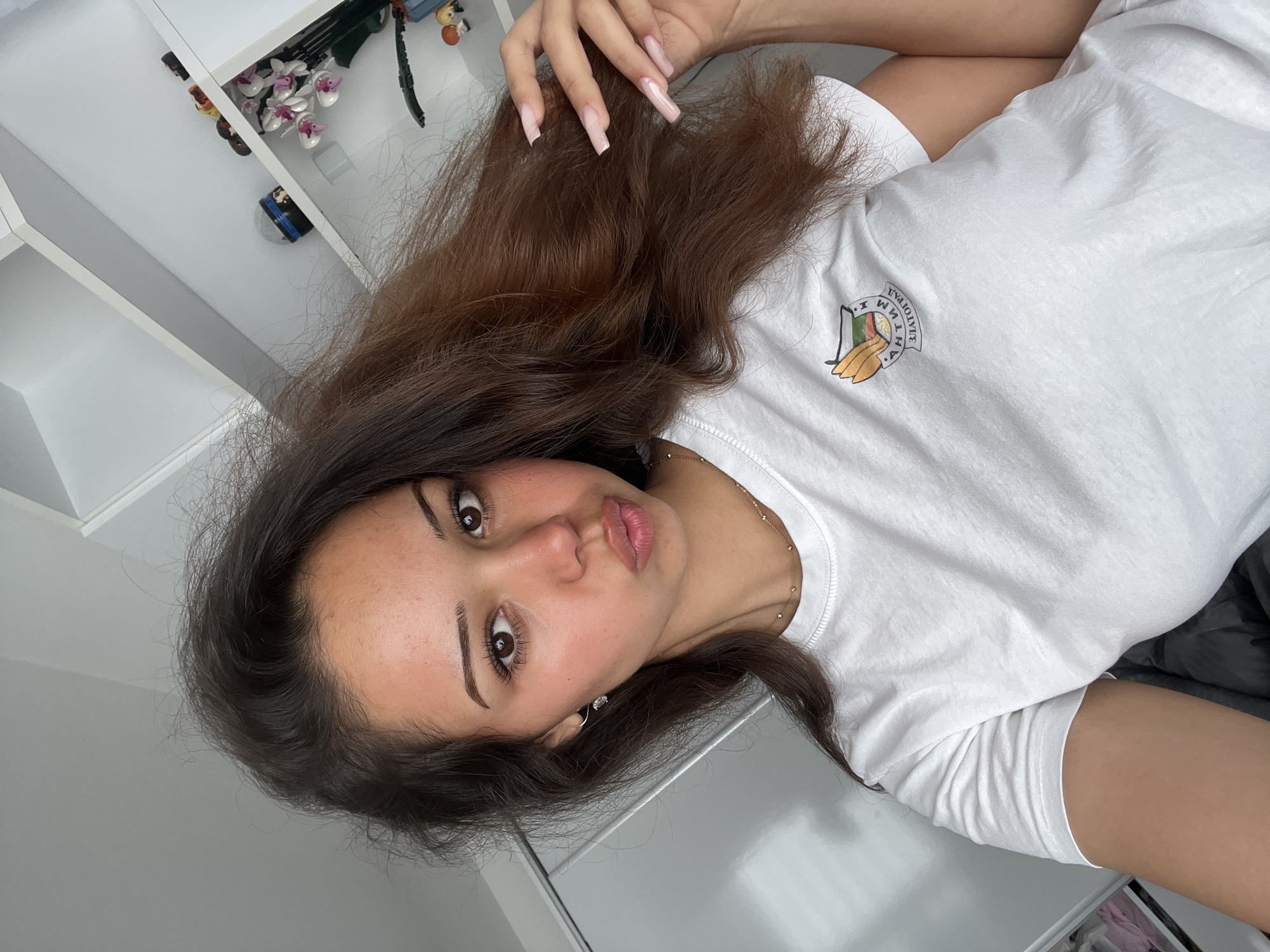 A young woman with brown hair, light skin, and brown eyes is posing for a selfie. She is wearing a white t-shirt with a crest on it that has Bulgarian text. The crest appears to be a coat of arms. She has on a small silver necklace and a hoop earring in her left ear. Her nails are long and painted a light pink. In the background, there are white shelves with various objects, including what appear to be decorative flowers and small figurines. The overall environment suggests an indoor setting, possibly a bedroom or living area, with bright lighting. The crest on her shirt includes text that says "АНТИМ" and "ЗЛАТОГРАД". Zlatograd is a town in the Smolyan Province of southern Bulgaria, known for its well-preserved Revival-era architecture and ethnographic traditions.