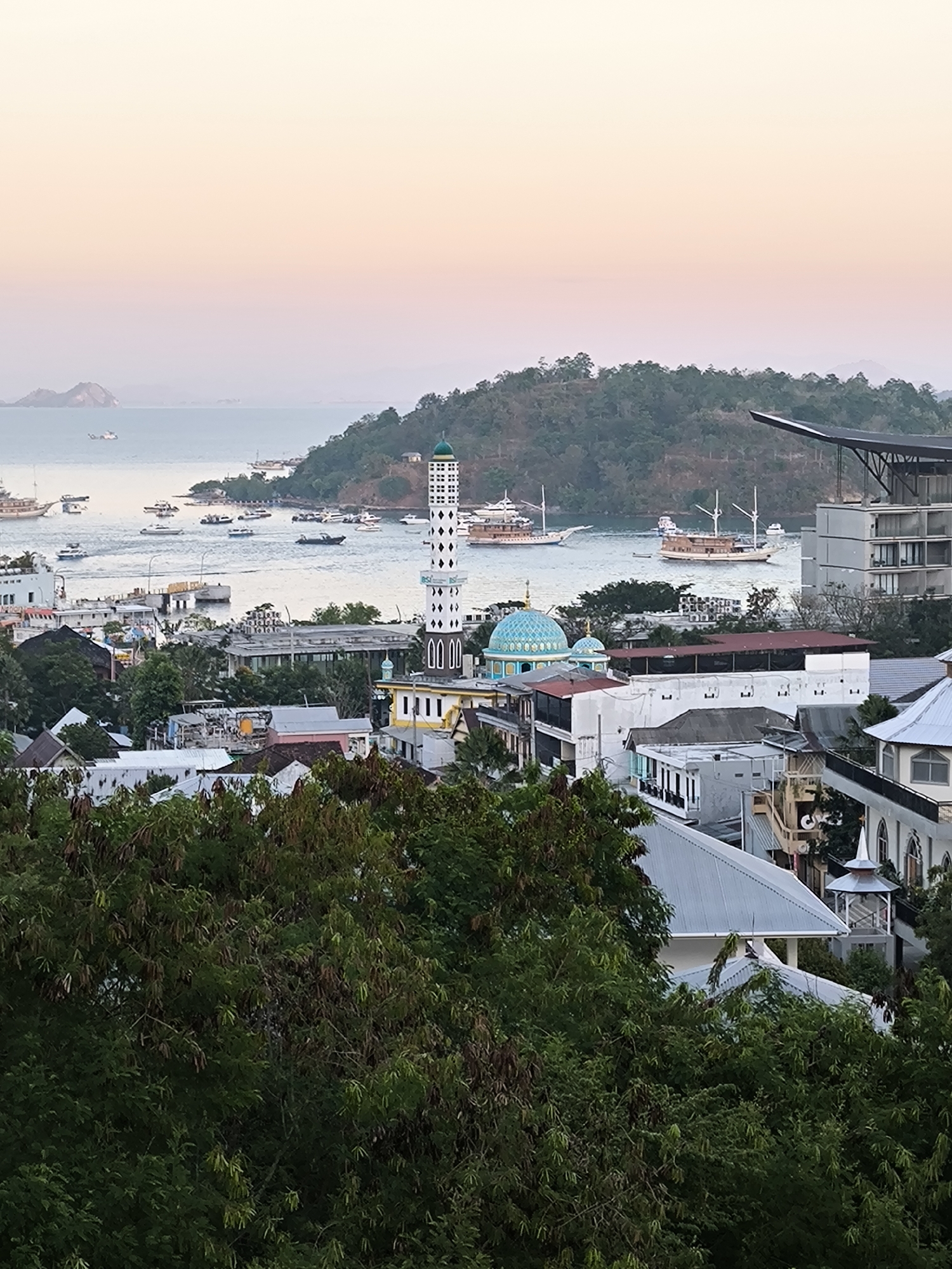This is a medium-angle, eye-level, outdoor, wide shot of a coastal city at sunset. The image depicts a harbor filled with boats, ranging from large sailing vessels to smaller motorboats. Buildings of varying architectural styles, including a mosque with a distinct blue dome and a tall minaret, are visible along the coastline. Trees and lush vegetation cover the foreground and the hills in the background. The sky is a gradient of soft orange and pink, indicating the approaching dusk. The scene suggests a bustling port town in a tropical climate. The presence of the mosque strongly suggests the location is in Indonesia, and given the harbor and overall aesthetic, it is likely the city of Labuan Bajo.