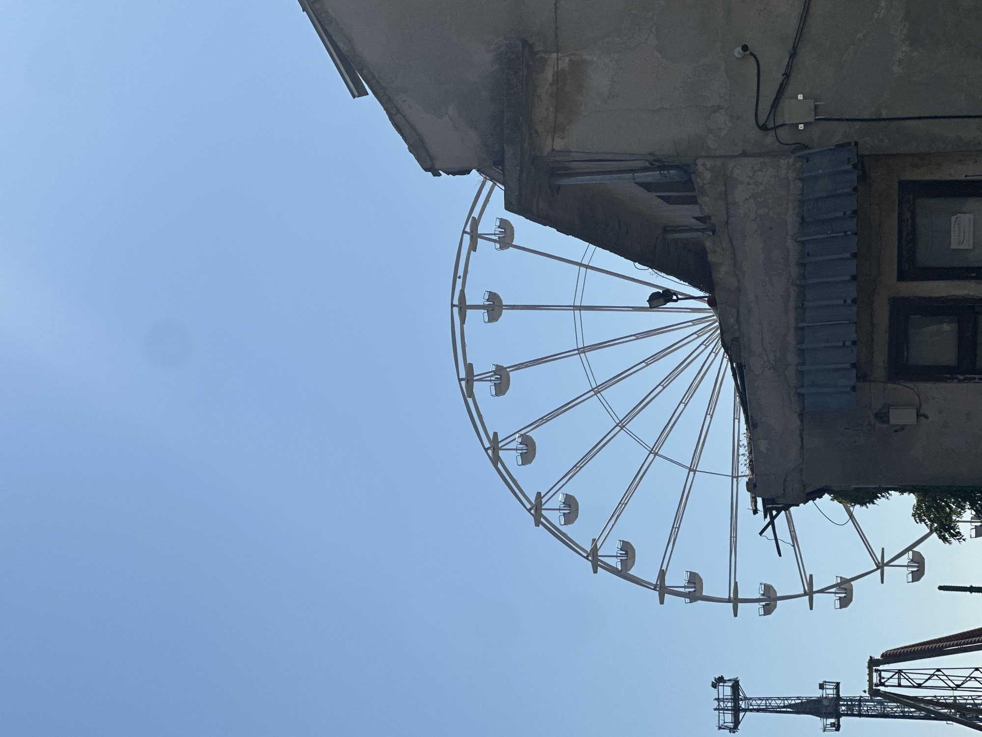 The original image shows a ferris wheel against a clear blue sky. In the foreground, a weathered, concrete building occupies the right side of the frame. The building has a damaged roofline and appears aged. To the left of the ferris wheel, a tall, metal tower structure, possibly part of a roller coaster or another amusement ride, is visible. The ferris wheel itself is a large, circular structure with spokes and multiple gondolas, though only a portion of it is in view. The overall scene suggests an amusement park or fairground setting during the day. There are no people visible in the image.