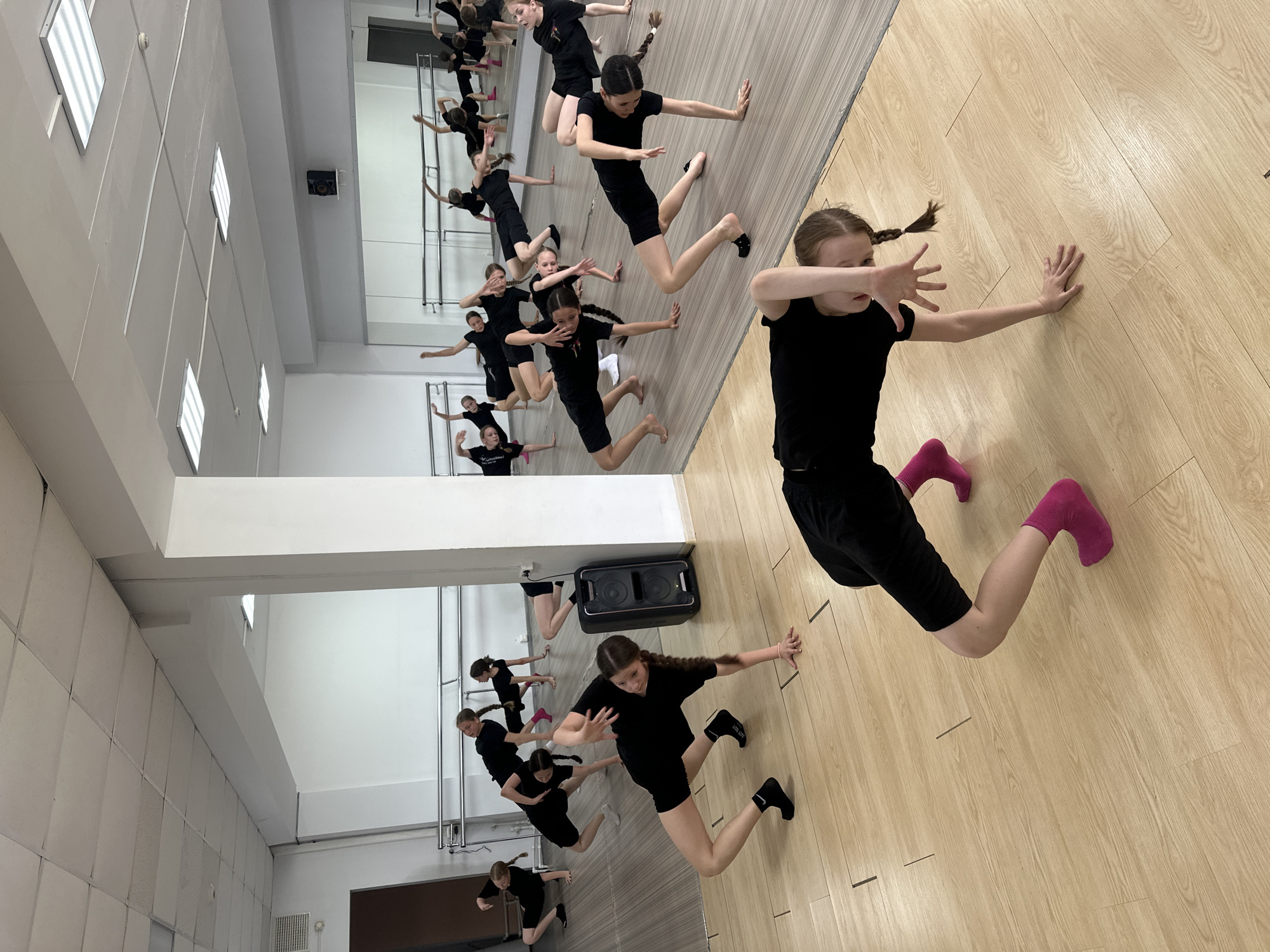In a dance studio with a light-colored wooden floor and white walls, a group of young girls in black athletic wear are practicing a dance routine. They are positioned in various dynamic poses, some on the floor with limbs extended, others in a low crouch. A large speaker sits against a white pillar on the right side of the frame. Mirrors line the back wall, reflecting the dancers and barre equipment. The ceiling is a white drop ceiling with rectangular light fixtures. The scene appears to be indoors, likely a dedicated dance or fitness space. The location in Votkinsk, Russia, is not discernible from the visual evidence in the image.