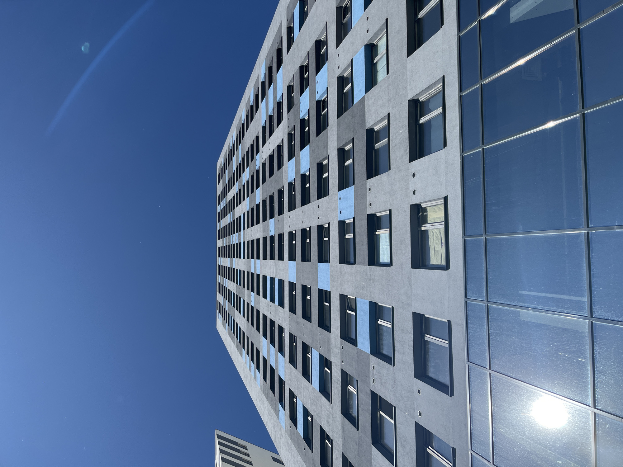 The image shows a low-angle view of a modern, tall, grey apartment building with blue rectangular accents on its facade. The building has a grid of uniformly spaced windows. The lower section of the building features a glass facade with reflections of the sky and sun. In the upper left corner, the edge of another building with vertical white and grey stripes is visible. The sky is a clear, deep blue, with a faint lens flare visible in the upper right quadrant, indicating a sunny day. The perspective emphasizes the height and scale of the building against the expansive sky. This building is located in the city of Saint Petersburg, Russia.