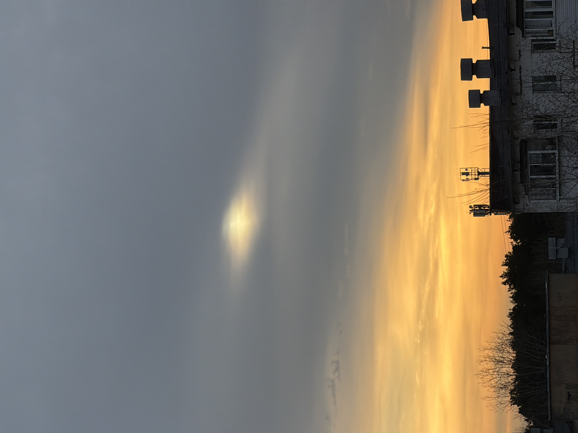The image depicts a cityscape, likely in Chaikovsky, Russia, during sunrise or sunset. The sky is divided into a grey, cloudy area on the left and a vibrant, orange-yellow area on the right. A faint, bright spot, possibly sunlight filtering through clouds, is visible in the grey area.
A tall building occupies the right side of the frame. It appears to be an apartment complex, based on the visible windows. The building's roof is lined with what appear to be ventilation shafts or other industrial-looking fixtures. The building is adjacent to a line of trees.