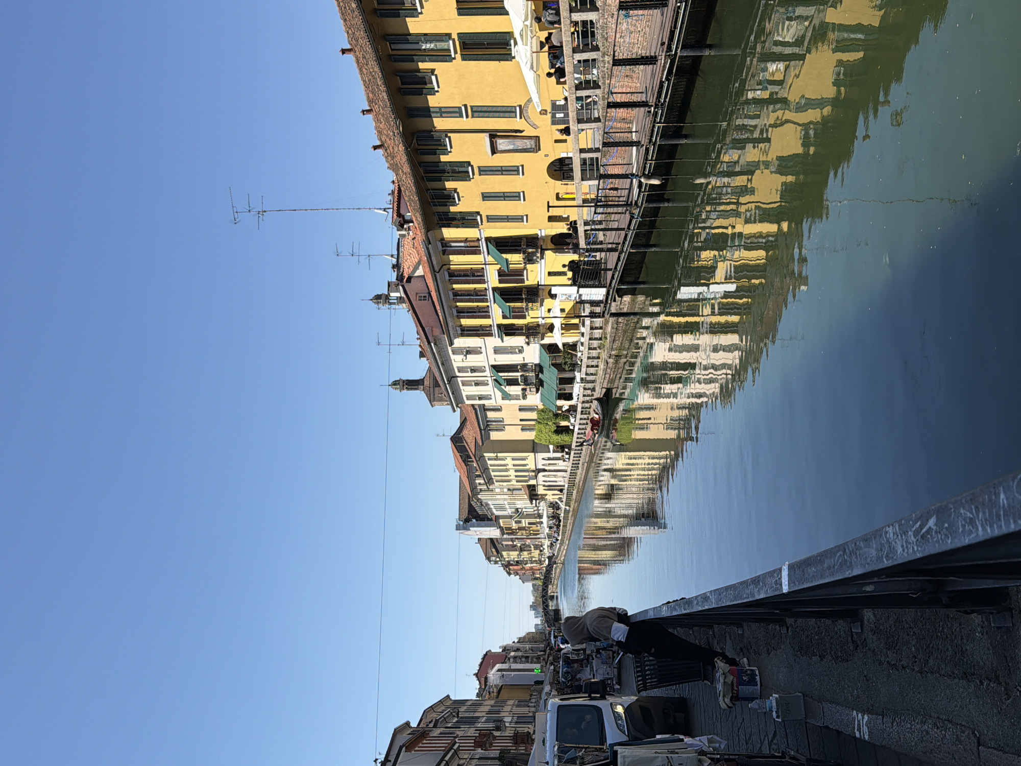 The image shows a canal in Milan, Italy, likely the Naviglio Grande, known for its historic canals and vibrant atmosphere. The canal reflects the colorful buildings lining its banks, which are a mix of yellow, beige, and other pastel hues. Some buildings have awnings and balconies, and there are antennas visible on rooftops. The water itself appears calm, creating a mirror-like effect for the architecture above. A railing runs along the canal's edge, suggesting a pedestrian walkway. There are parked vehicles, including a white van, visible on the street beside the canal. There are people walking and sitting near the canal.