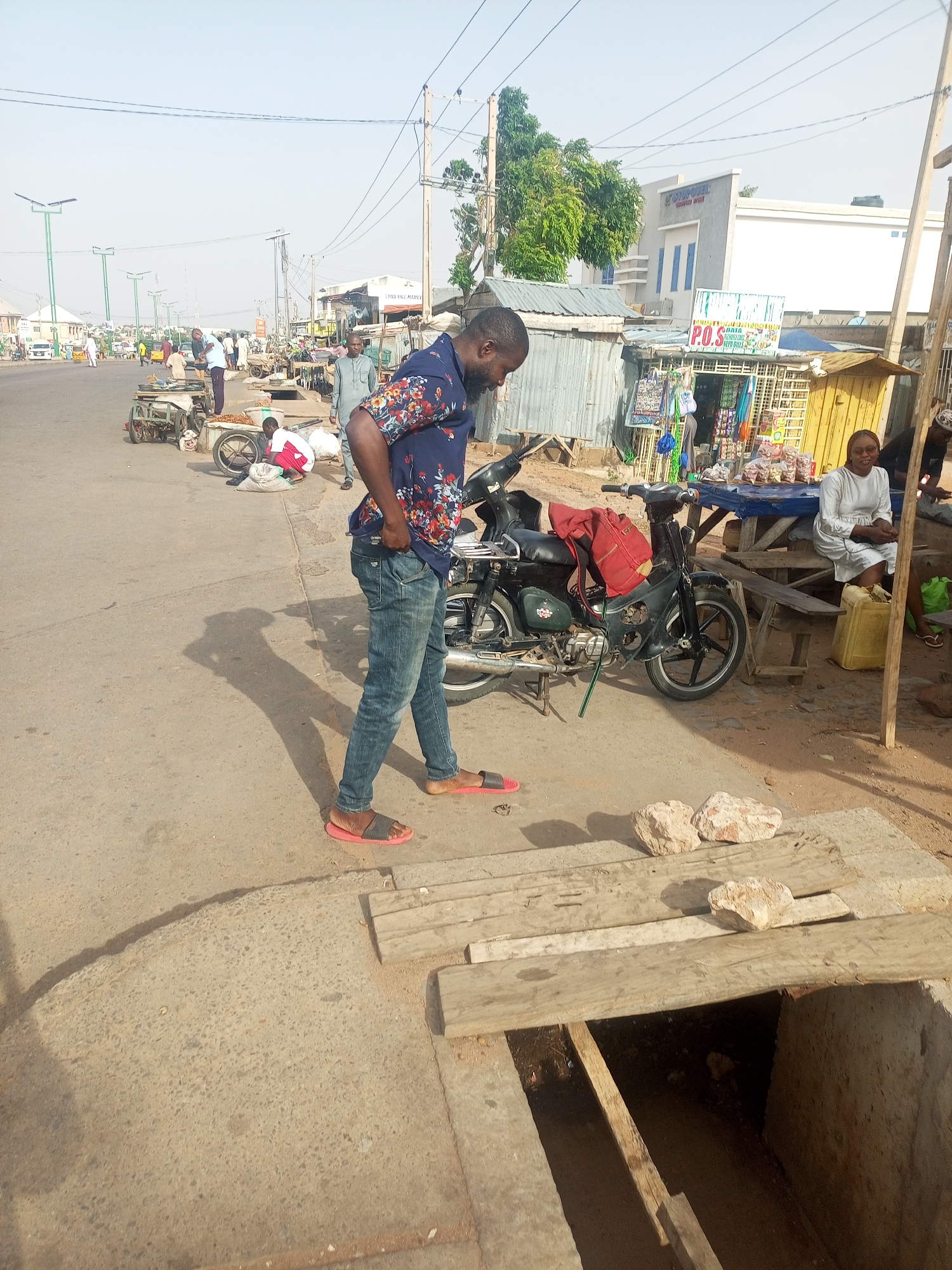 The image depicts an urban scene in Tempure, Nigeria, focusing on a section of a street.
A man in a blue patterned shirt and jeans stands near a parked motorcycle on a paved surface. He is looking down, and his left hand is in his pocket. His shadow stretches across the pavement.
Near him is an open concrete drain covered partially with wooden planks and secured with rocks.
In the background, there are other people, market stalls, and buildings typical of a busy street. Some people are sitting, and others are moving around. Vehicles are visible further down the street, and overhead power lines are strung across the scene.