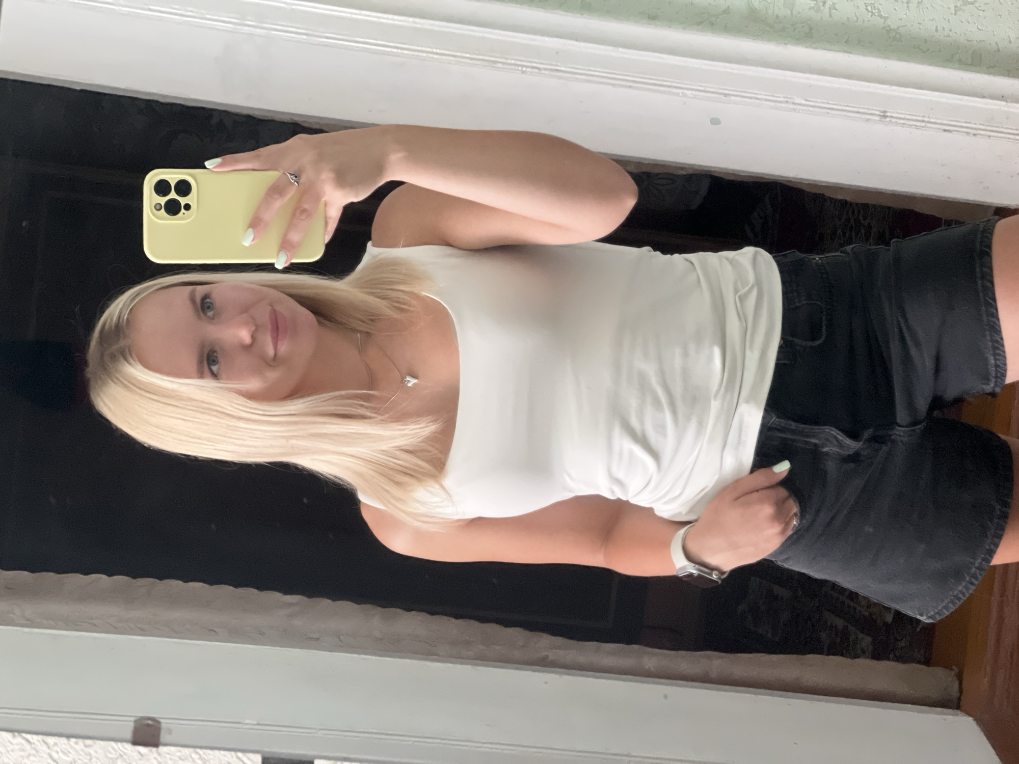 In this medium shot, a young Caucasian woman with blonde hair and blue eyes is captured in a mirror selfie. She is wearing a white tank top and black denim shorts. A pale yellow smartphone is held in her right hand, and her left hand is in her pocket. She is also wearing a silver necklace with a heart-shaped pendant and a silver ring on her left hand. A white smartwatch is visible on her left wrist. The background appears to be an indoor setting, possibly a room with a doorway and a mirror. The lighting is natural, coming from the front.