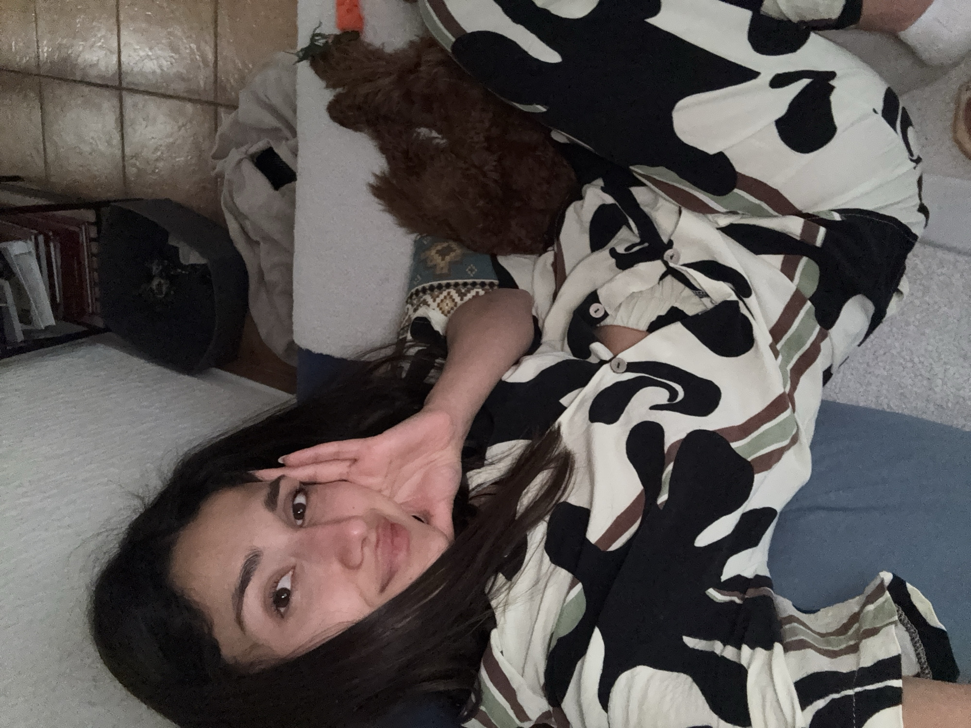 A young woman with long dark hair is lying on a blue surface, possibly a bed or sofa. She is wearing patterned pajamas with a print in shades of black, white, brown, and green. Her left hand is raised, partially covering her face in a gesture. Behind her, on a white surface, is a brown stuffed animal. In the background, on the left, is a shelf with books and a black object. The location of the photo is not discernible to be Sutomore, Montenegro.