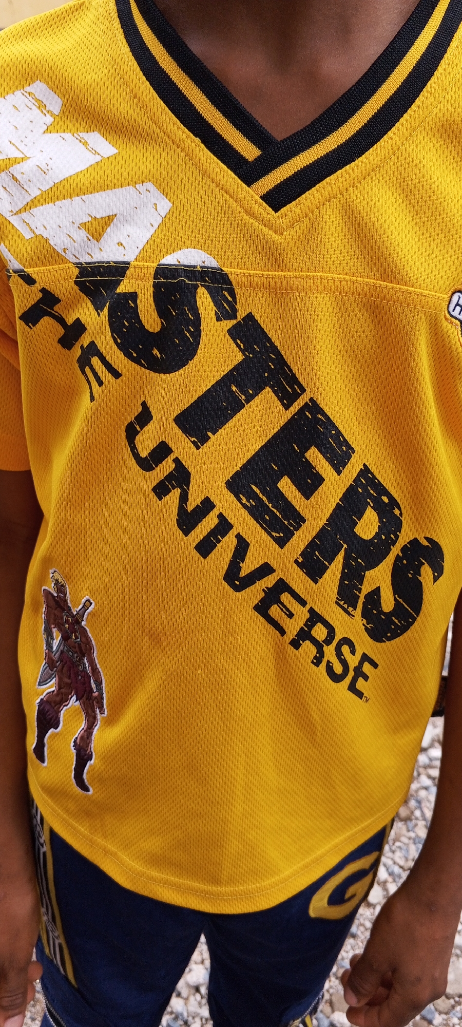 A close-up, eye-level, outdoor shot captures the torso of a young individual wearing a yellow, mesh jersey with black and yellow striped trim around the V-neck. Bold, black, distressed lettering on the jersey reads "STERS UNIVERSE." A small, full-body illustration of a cartoon warrior figure, possibly He-Man, is affixed to the left side of the jersey. The jersey is paired with dark blue shorts featuring a yellow stripe design. The background consists of a gravelly surface. The scene appears to be an informal outdoor setting, possibly a residential area or a public space in Jalingo, Nigeria.