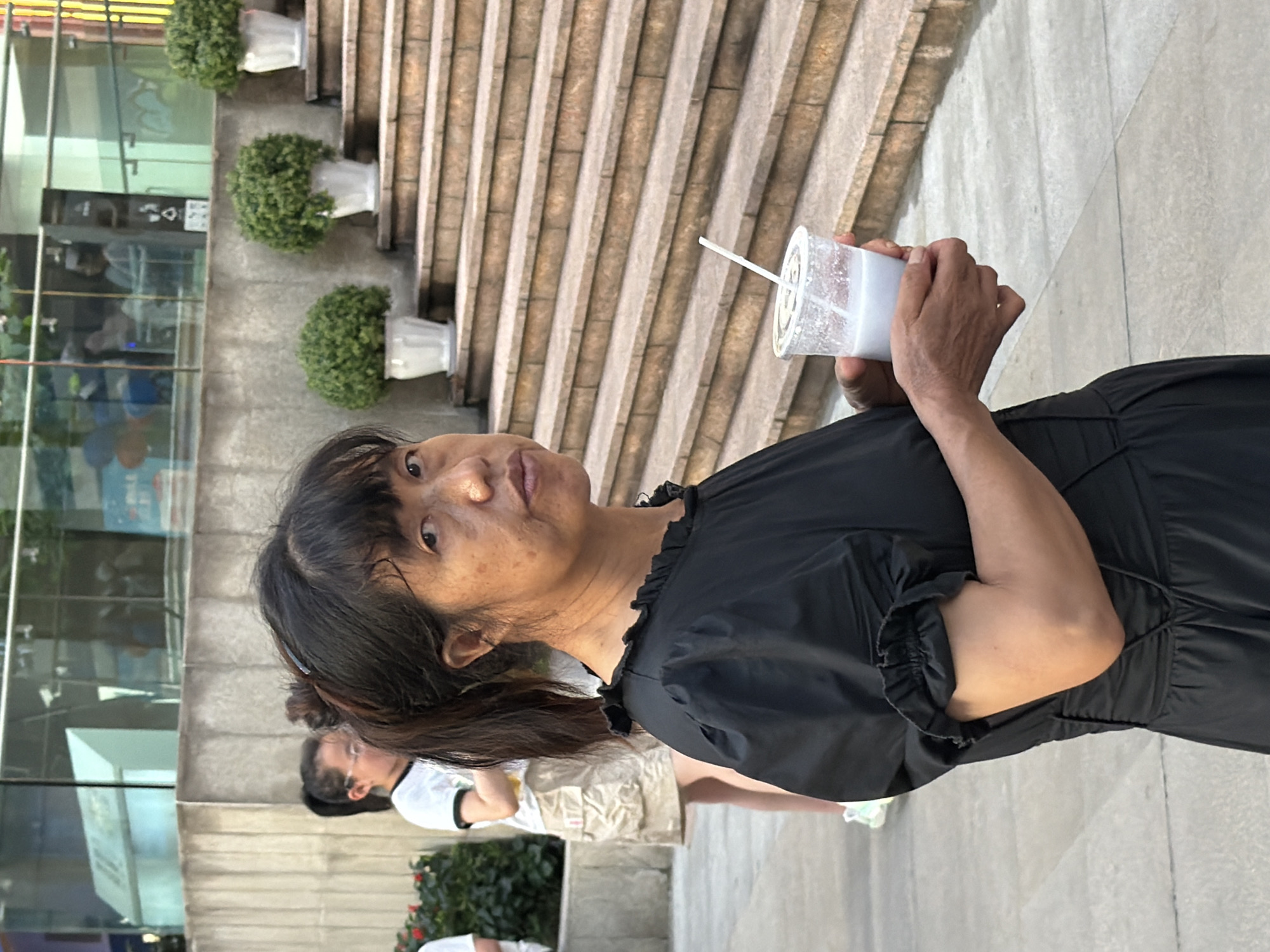 The image shows a woman in a black dress holding a clear plastic cup with a white lid and a straw, containing a milky white beverage. She is looking towards the camera with a neutral expression. Her hair is dark and pulled back into a ponytail. In the background, there are several potted green plants placed on ascending steps. To the left of the woman, there is a glass building with reflections of people and signage. Another person, wearing a white t-shirt and shorts, is visible in the background, partially obscured by the main subject. The overall setting appears to be an outdoor urban environment, possibly a commercial area, given the presence of the glass building and the structured steps with planters. The lighting suggests it is daytime, and the weather appears to be pleasant. No visible text is discernible in the image.