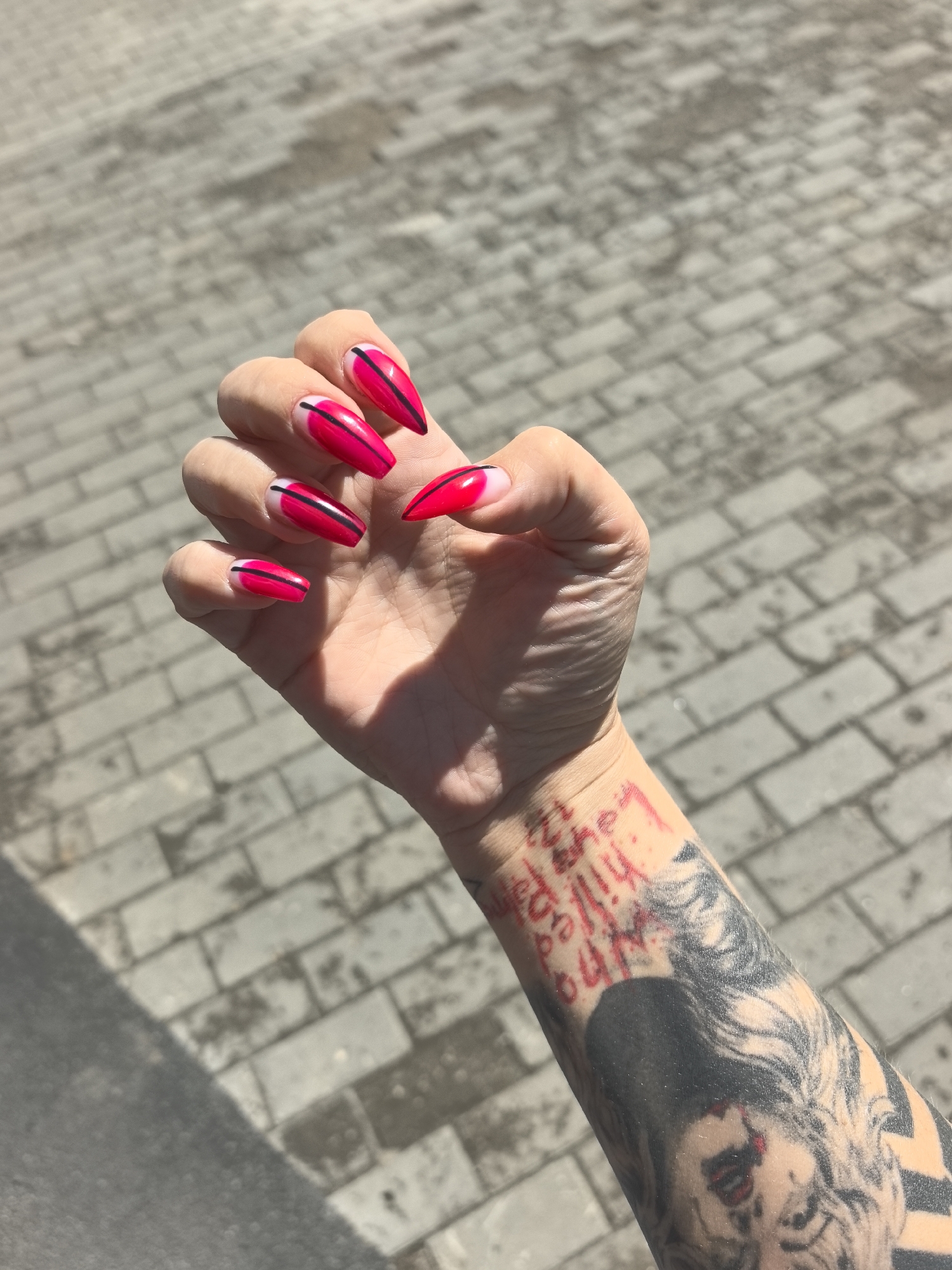 A close-up, high-angle shot captures a person's left hand and forearm extended over a paved ground in Chaikovsky, Russia, under bright daylight. The hand showcases long, coffin-shaped nails painted in a vibrant hot pink (fuchsia) color with a distinct black horizontal stripe across each nail, set against a natural or clear base near the cuticles. The fingers are slightly splayed, displaying the nail art.
The forearm is heavily tattooed. On the inner wrist, red script text is visible, which appears to read "I will never give up", "I will never lose", and "i know that", though some parts are partially obscured and stylized. Below this text, a large black and white tattoo of a person's face is prominent, featuring detailed shading and lines, though the subject of the portrait is not clearly identifiable.
The background consists of light gray rectangular paving stones or cobblestones, suggesting an outdoor urban or suburban setting. The bright sunlight casts distinct shadows, indicating a clear, sunny day, likely around midday. The image focuses on personal style through the nail art and tattoos.