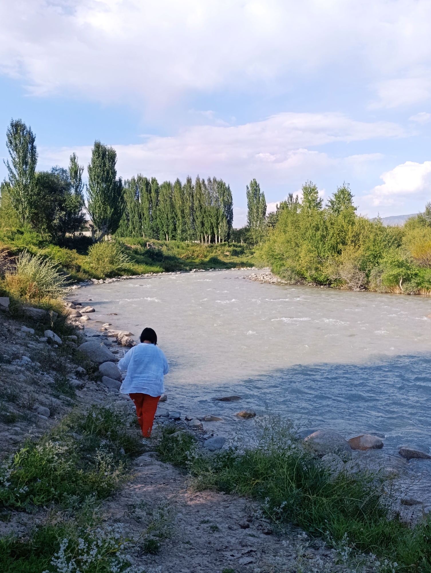 A person with dark hair wearing a light blue shirt and orange pants is walking on a rocky, grassy path next to a river in Kyzyl-Jyldyz, Kyrgyzstan. The river is wide and has murky, rippling water. The path is uneven and strewn with stones. Lush green trees and vegetation line the opposite bank of the river, with a few scattered trees also visible on the near bank. The sky above is partly cloudy with patches of blue.