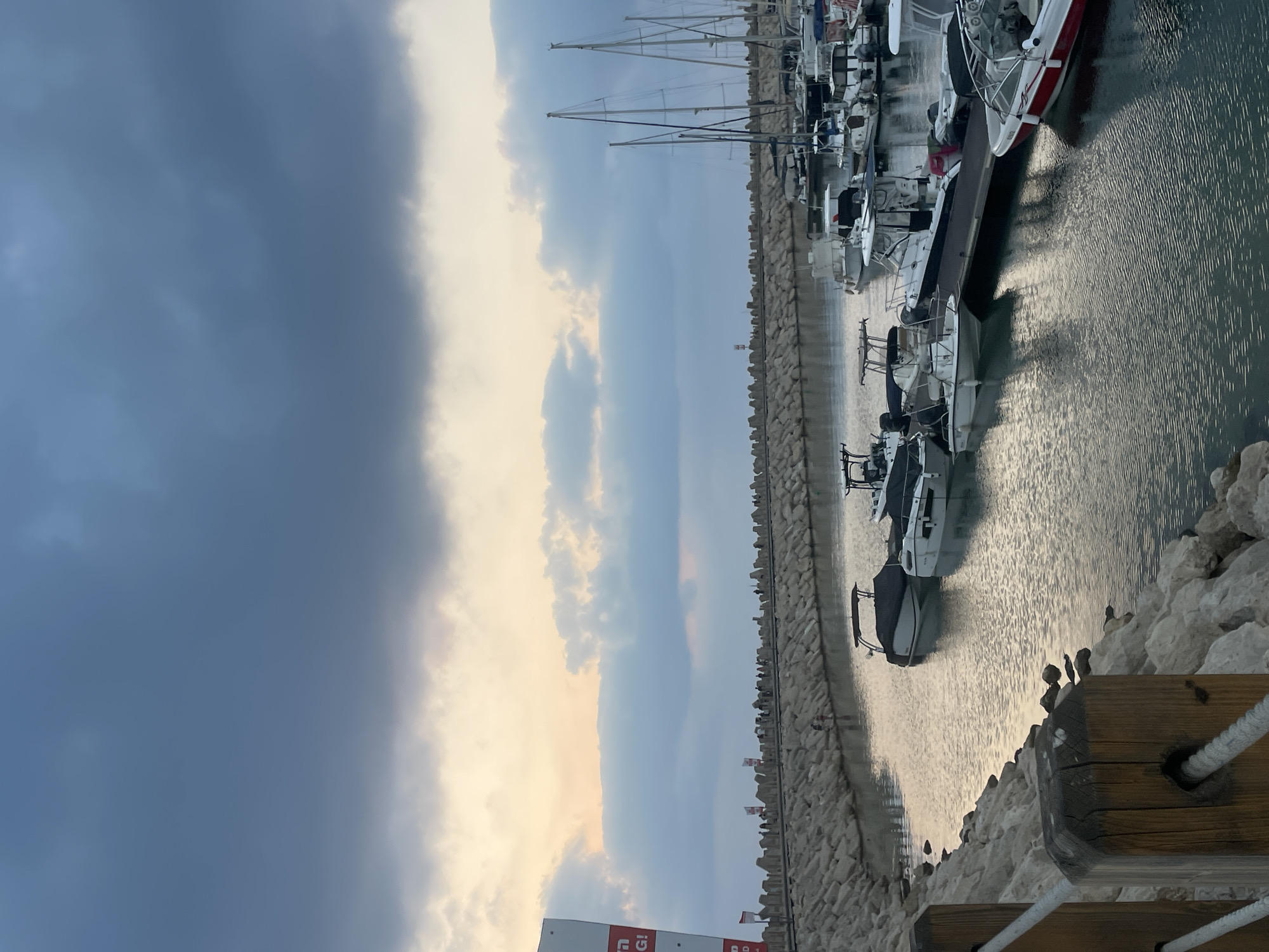 The image captures a serene marina in Herzliya, Israel, under a cloudy sky. The water in the marina is calm, reflecting the muted tones of the sky. Several boats, ranging from small motorboats to sailboats, are docked and moored, with some covered and others exposed. A stone breakwater, lined with people walking along its top, separates the marina from the open sea. The breakwater is constructed from large, irregularly shaped stones, creating a rugged barrier. On the left side of the image, a large white sign with Hebrew text is partially visible, indicating a commercial or informational presence. The overall atmosphere is tranquil, suggesting either early morning or late evening light.