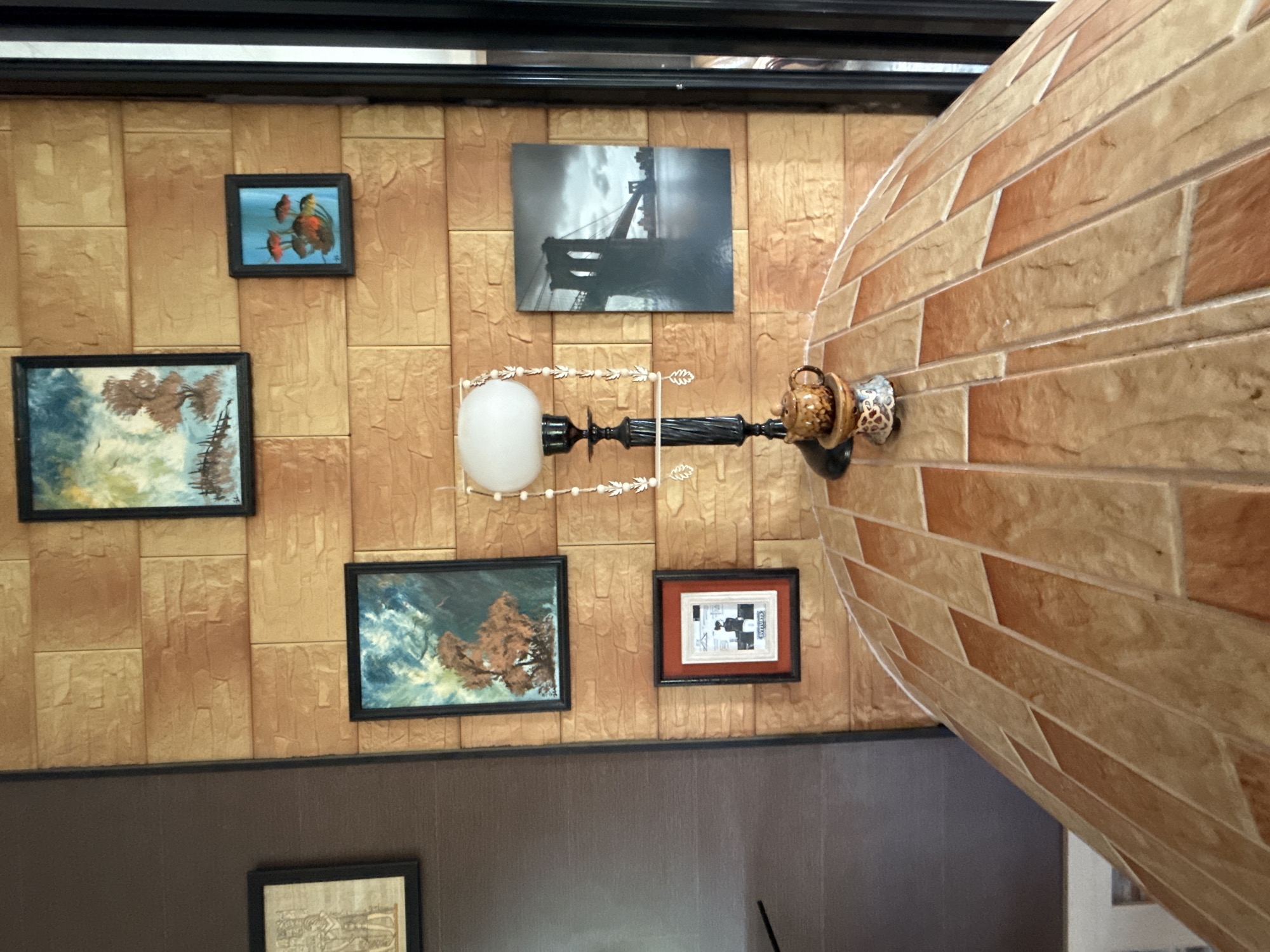 The image shows a close-up of a lamp on a tiled surface, with a wall adorned with several framed artworks behind it. The wall has a textured, faux-brick finish in shades of tan and orange.
On the wall, from left to right and top to bottom, there are:
- A framed piece of art featuring figures that appear to be from an ancient civilization.
- A landscape painting with a tree and a fence, possibly depicting a winter scene with blue and green hues.
- A small framed artwork showing what looks like a person in silhouette with text below, possibly "GVITTURYS".
- A small framed painting with red and orange shapes against a blue background, resembling possibly fish or coral.
- A large black and white photograph of the Brooklyn Bridge under a cloudy sky.
The lamp has a white globe shade and a decorative, twisted black base with hanging beaded ornaments. Below the lamp, on the tiled surface, sits a small, ornate ceramic teapot. The tiled surface is curved and made up of rectangular tiles in alternating shades of light tan and reddish-brown. The image is taken indoors, in what appears to be a room with a warm, decorative aesthetic. The specific location in the city of Galinė, Lithuania is not discernible from the image content.