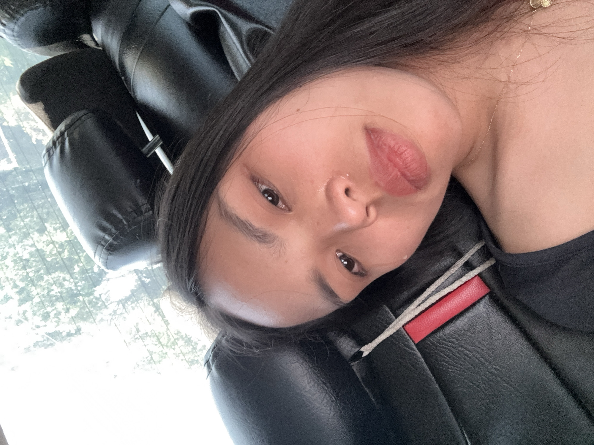 A close-up, angled photograph depicts a person with light skin and dark hair, likely female, reclining against a black leather car seat. The person faces the camera with a slightly downturned gaze. A visible gold necklace is worn around the neck. Part of a white seatbelt and red accent are visible on the seat. The background shows glimpses of foliage and daylight, suggesting the vehicle is moving or parked outdoors. There is no specific evidence within the image to confirm the location in Davao City, Philippines.
