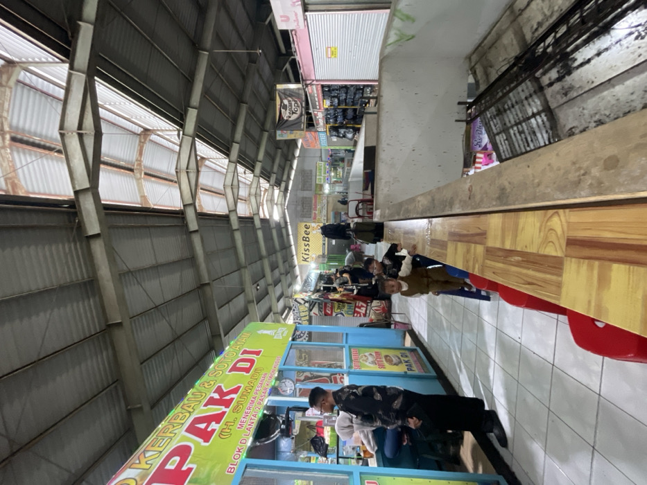 This image portrays an indoor market or food court area in Nganguk, Indonesia. The overhead structure consists of a corrugated metal roof with exposed steel truss work and multiple translucent skylight sections, providing natural illumination supplemented by artificial lights from various stalls.

In the left foreground, a food stall named "PAK DI" is visible, featuring a green and yellow sign advertising "SOTO KERBAU & SOTO AYAM." The sign specifies "BLOK D LANTAI 2 PASAR KLIWON KUDUS" as its location and indicates it accepts orders. The stall has blue-framed window sections and a counter. An adult male, wearing a black patterned long-sleeved shirt and dark trousers, stands facing left in front of the stall. Behind him, another individual, partially obscured and wearing a light-colored hijab, is seated on a blue stool at the stall's counter.

To the right, a wooden-patterned countertop extends horizontally, acting as a barrier. Two young children stand at this counter. A toddler, dressed in a brown vest over a long-sleeved shirt and brown trousers, stands on a blue plastic stool, resting a hand on the counter. Adjacent to this child, a slightly older child wearing a dark top and glasses also stands at the counter. Red plastic stools are visible below the counter on the left.

The wooden-patterned counter overlooks a lower level that appears to be unfinished or disused, characterized by exposed concrete walls, steel rebar, and a metal grate. A partially visible sign, "Sis-Dh...", is located within this lower section.

Further into the background, numerous other market stalls are present. One stall prominently displays a "KissBee" sign, while another features a black banner with text, possibly "HEROON TOPI." Several stalls exhibit merchandise, and some have closed metal roller shutters. The floor in the immediate foreground consists of white square tiles.