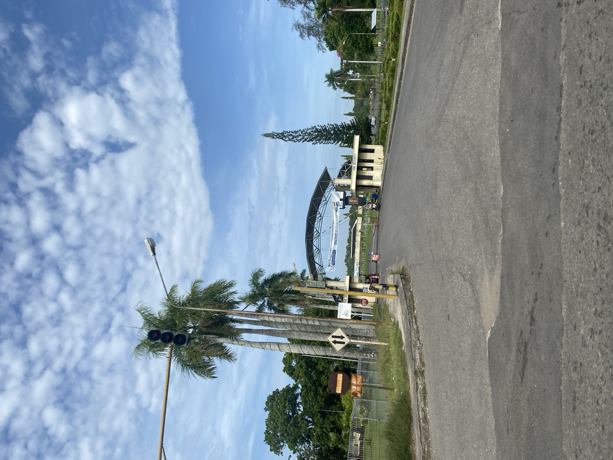 The image shows a view of a road in Lhokseumawe, Indonesia. The road appears to be paved with asphalt. A traffic light is positioned on the left side of the road. Palm trees line the side of the road near the traffic light. A sign indicates that there are two possible directions for traffic. In the distance, there is a structure that resembles a gate or entrance to a facility. The sky is blue with white clouds. The overall scene suggests a sunny day in a tropical environment. There are people riding motorbikes near the gate.