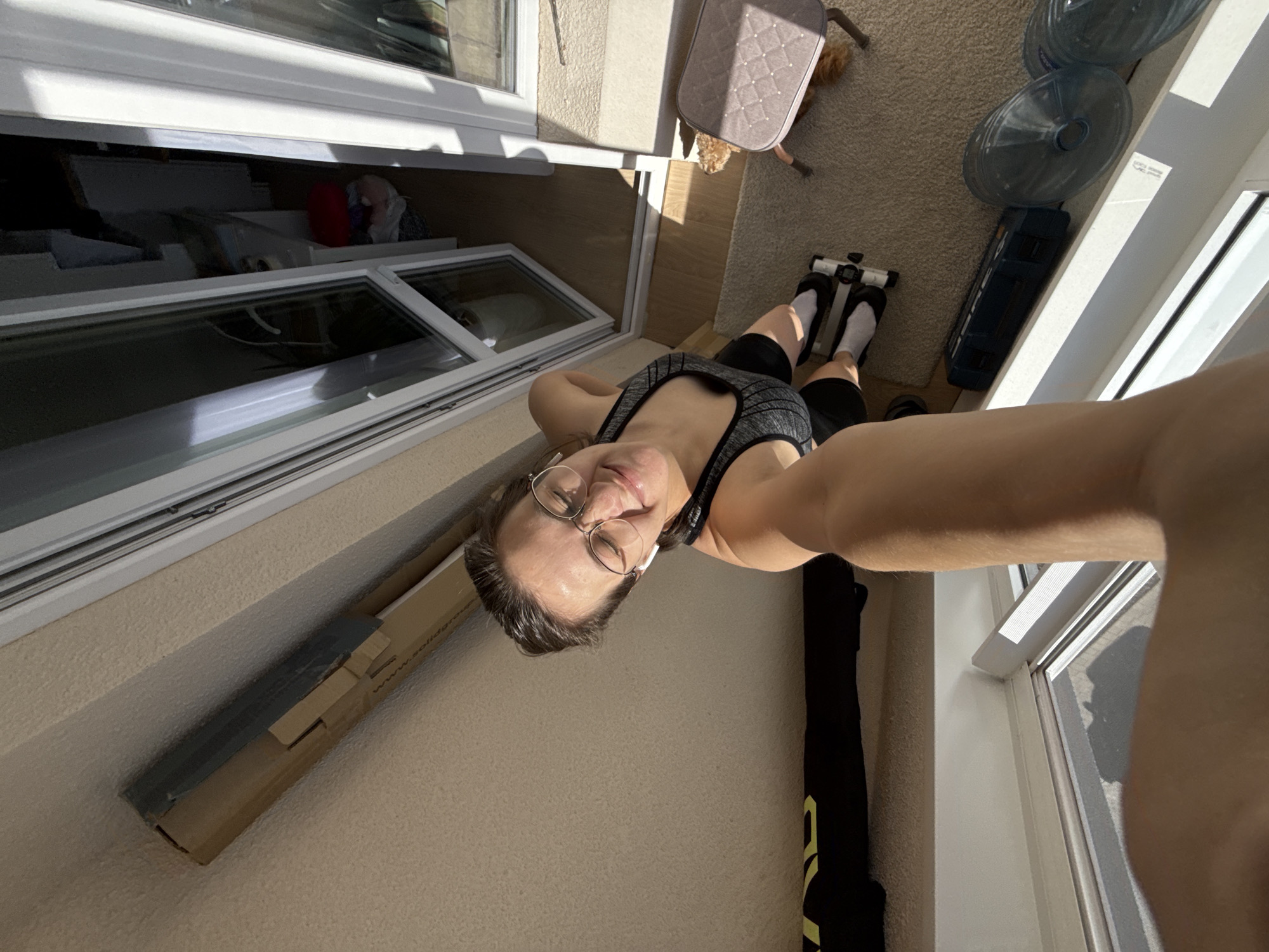 A woman is captured in a high-angle selfie, looking down into what appears to be a sunlit indoor space in Pervomaysky, Russia. She is smiling pleasantly, wearing round glasses, a grey patterned sports bra, and dark shorts. Her feet, clad in white socks, are positioned on a small piece of exercise equipment, possibly a foot pedal exerciser or an ab roller.
The setting features light beige carpeting on one side, where two large clear water bottles and a dark blue rectangular hard case are placed. A small, grey patterned pet carrier or bed is also visible on the carpet, with a light-colored dog peeking out from underneath. The room is brightly illuminated by direct sunlight, casting strong shadows, indicating it is daytime.
Two white-framed windows dominate the left side of the image. The lower window is slightly ajar, revealing a darker, perhaps recessed, area behind it. Adjacent to the woman, against a textured beige wall or floor, is a cardboard box with partially visible text "www.solidg". A black bag or cover, possibly for sports equipment, with some yellow detailing, lies near her arm. The overall scene suggests a relaxed moment in a personal fitness or living area.