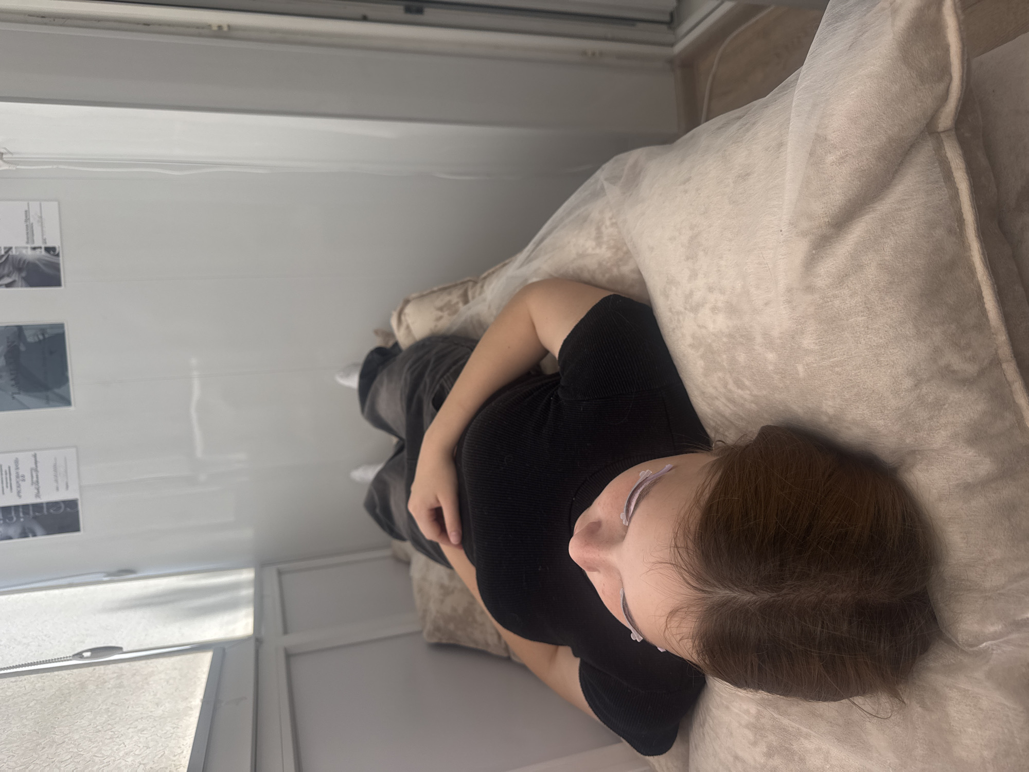 The image shows a person lying on a cushioned surface, possibly a bed or massage table. The person is wearing a black top and dark pants. The environment appears to be an indoor space with white walls, and a window with blinds is visible on the left. There are some framed items on the wall in the background. The person's eyes are closed and there appear to be some materials applied to their eyelashes. The scene suggests a beauty or spa treatment is taking place. The location is identified as Saratov, Russia.