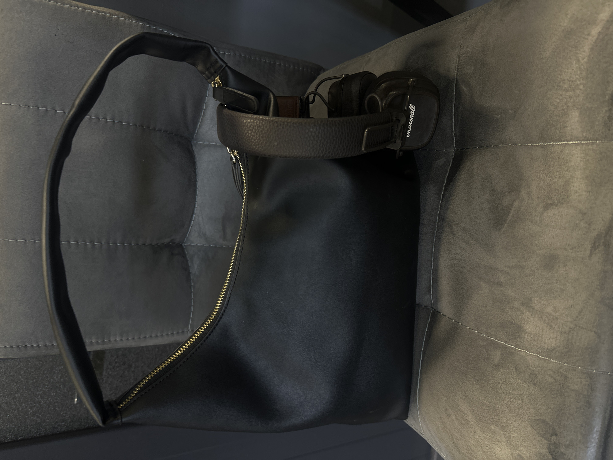 The image shows a black shoulder bag sitting on a gray cushioned chair. A black pair of Marshall brand headphones rests on the bag's handle. The bag has a gold zipper. The chair appears to be indoors, possibly in a domestic setting. There is no indication of Yerevan or Armenia in the image.