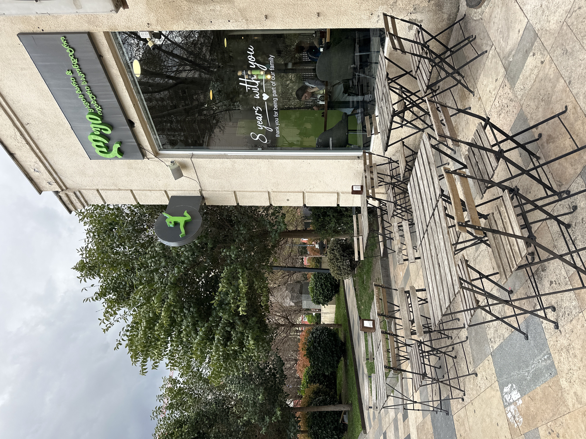 The scene captures an outdoor seating area of a café in Tbilisi, Georgia. The façade of the café is light beige with a green sign in Georgian language along the top and a smaller green logo with a dog-like figure. The window of the café bears the inscription "8 years with you," a thank you note for customers being part of the establishment's family. Inside, a woman is seated at a table.
Several wooden tables and metal chairs are arranged in rows on a beige tile surface. Beyond the outdoor seating, a garden with green trees and bushes extends into the background. The sky above is overcast. The photo is taken from an angled, ground-level perspective, which slightly rotates the entire scene.