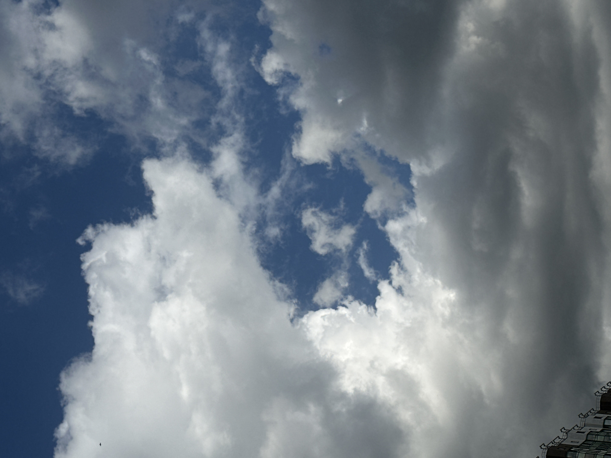 This is an outdoor, eye-level, medium shot photograph of a cloudy sky with a section of a building visible in the lower left corner. The sky is a deep blue, with a mix of white and gray clouds scattered throughout. Some of the clouds are dense and dark, while others are light and fluffy. A small, dark object, possibly an airplane, is visible in the upper left quadrant of the image against a white cloud. The building appears to be a multi-story structure with a modern architectural design, featuring glass windows and a visible roofline with decorative elements. The overall lighting suggests it is daytime.