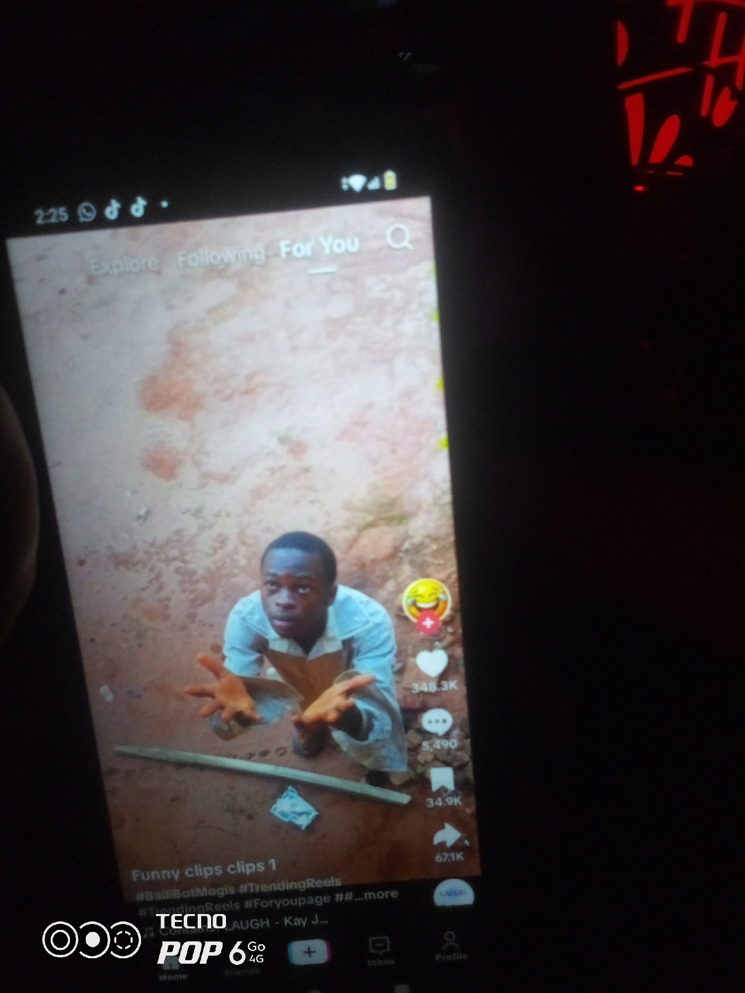 The image displays a smartphone screen showing a social media video. The video features a young Black male, appearing to be in his late teens or early twenties, looking upwards with his hands outstretched. He is wearing a light blue collared shirt and is positioned in a dirt-like environment. On the right side of the screen, social media engagement metrics are visible, including likes, comments, shares, and views. At the bottom of the screen, a navigation bar indicates various options like "Home," "Friends," and "Profile," along with the "TECNO POP 6 4G" branding. The top left corner shows the time "2:25" and icons representing WhatsApp, TikTok, and possibly another app. The background of the video appears to be a textured, earthy surface, potentially a wall or ground. The image is taken from an angled perspective, with the right side of the smartphone screen partially obscured by darkness. There is no visible information in the image that suggests a specific location in the city of Howayi, Nigeria.