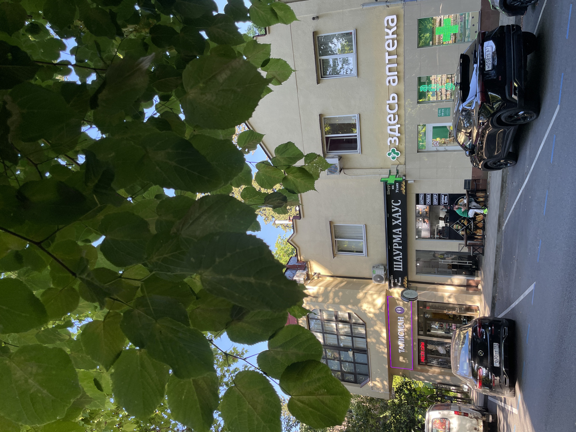 A vertical, low-angle outdoor shot shows the facade of a building with shops on the ground floor, partially obscured by large green leaves in the foreground. The building is cream-colored, with windows visible on the upper floors. One prominent sign reads "здесь аптека" (here is a pharmacy) in large white lettering, with a green cross symbol. Below this, another sign says "ШАУРМА ХАУС" (Shawarma House), indicating a food establishment. A black car is parked on the street in front of the shops, with its license plate visible. Another white van is parked further down. The scene appears to be a street in a town or city, possibly during daylight hours judging by the lighting and shadows. The overall impression is that of a commercial street with a focus on everyday services.