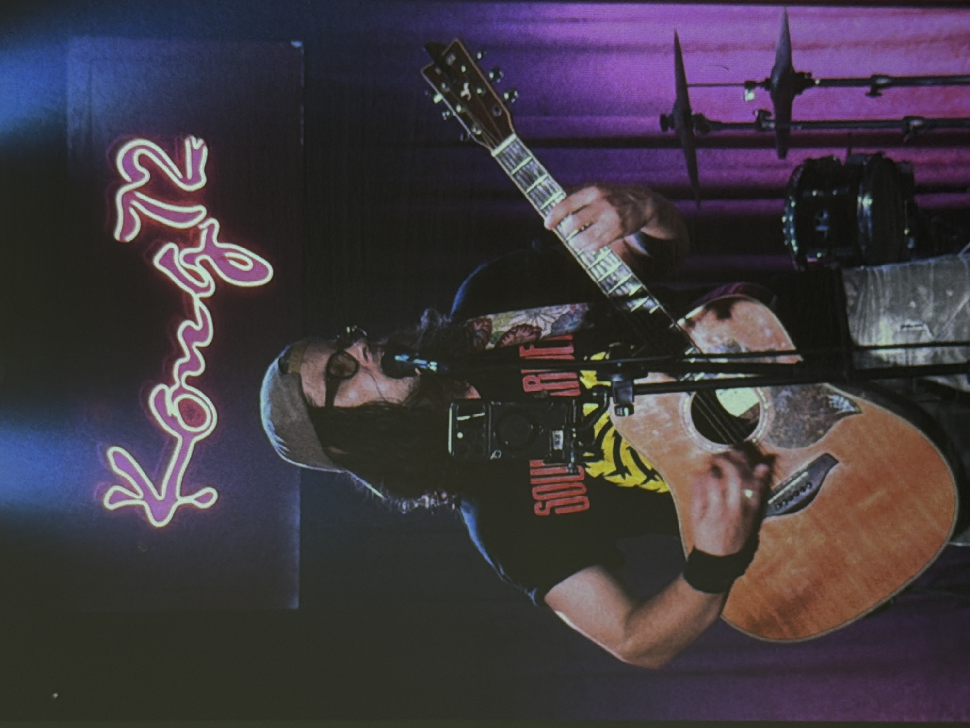 A man with long hair, wearing glasses and a hat, is playing an acoustic guitar on a stage in front of a purple curtain and drum set. He is wearing a black t-shirt with red and yellow lettering. A neon sign resembling stylized text, possibly in Vietnamese, hangs to the left of the stage. The sign is purple and white. The location is likely a music venue, although the specific location within Đà Nẵng, Vietnam is not identifiable from the image.