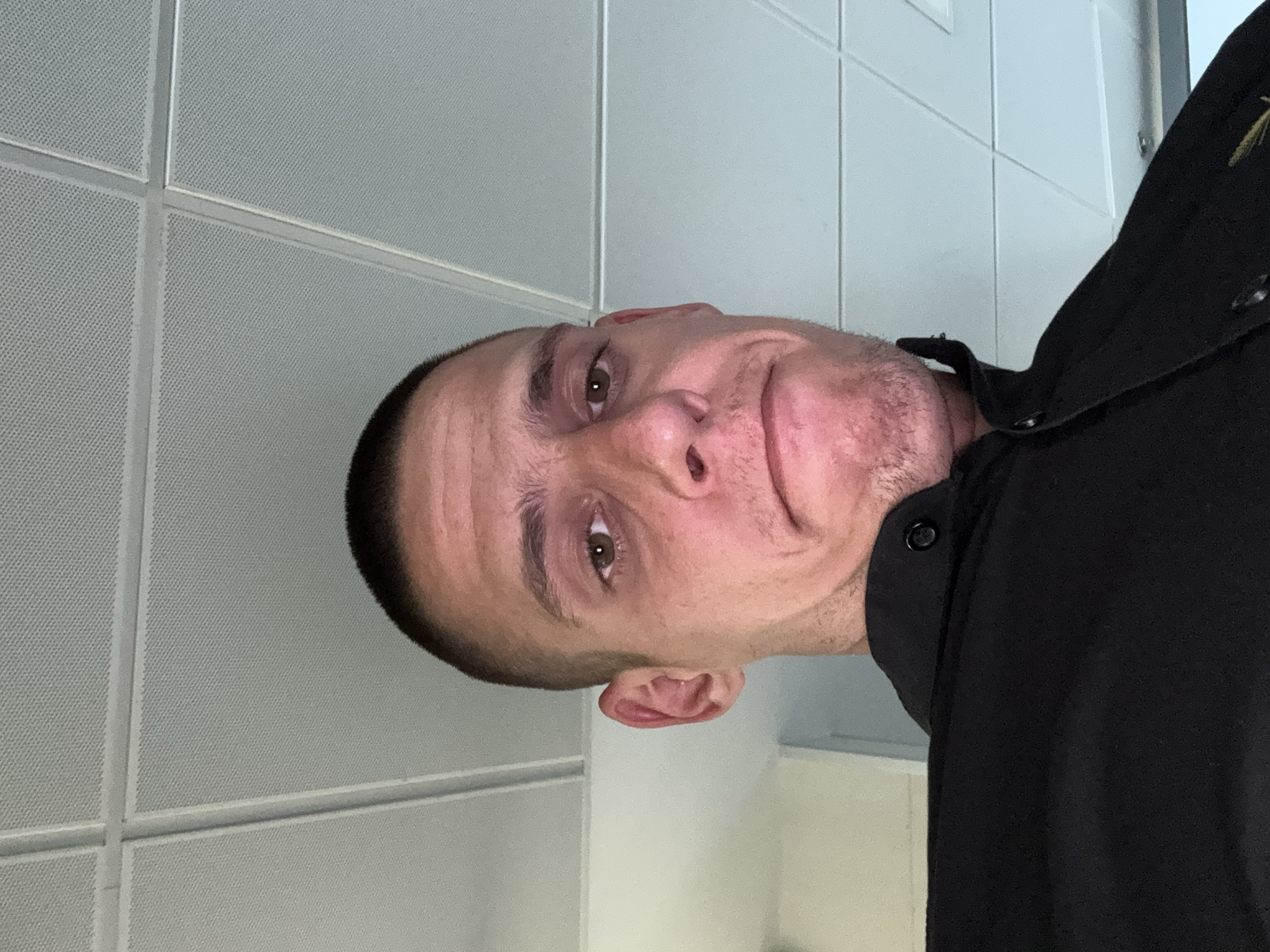 A close-up, head-and-shoulders shot of a light-skinned man with a shaved head, wearing a black button-down shirt. The background consists of a white, tiled ceiling and wall. The man's expression is neutral. The location cannot be determined from the image. There is no verifiable indication of the image being taken in Sofia, Bulgaria.