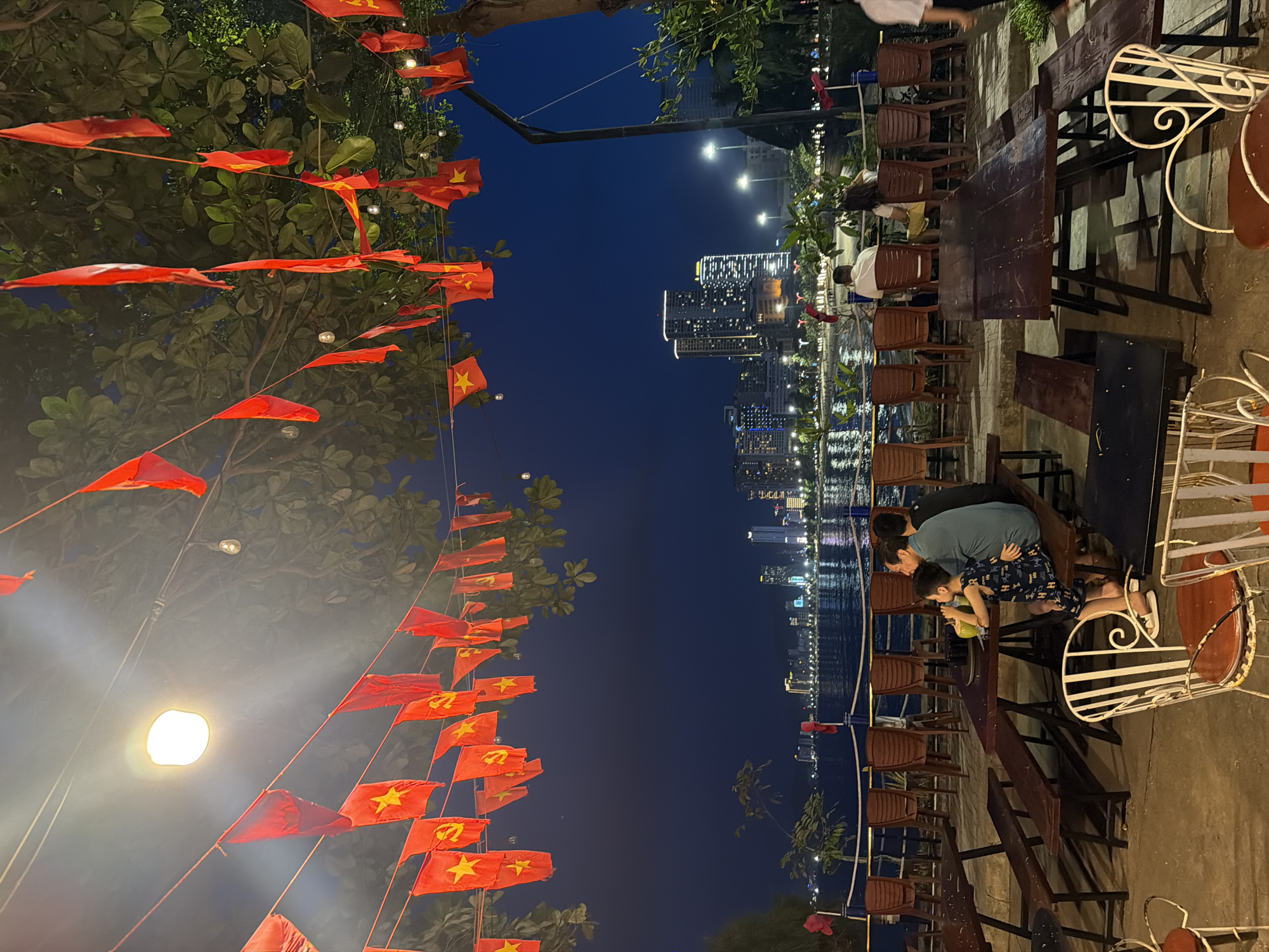 In Nha Trang Ward, Vietnam, at night, rows of red flags with yellow stars, some with hammers and sickles, are strung across tree branches over an outdoor seating area. Several wooden picnic tables and benches are arranged on a paved surface. In the background, a body of water reflects the lights of a city skyline with numerous high-rise buildings. A family of four, including two adults and two children, are seated at one of the tables, with one child holding a drink. The scene suggests a casual dining or gathering spot with a patriotic or festive atmosphere.
