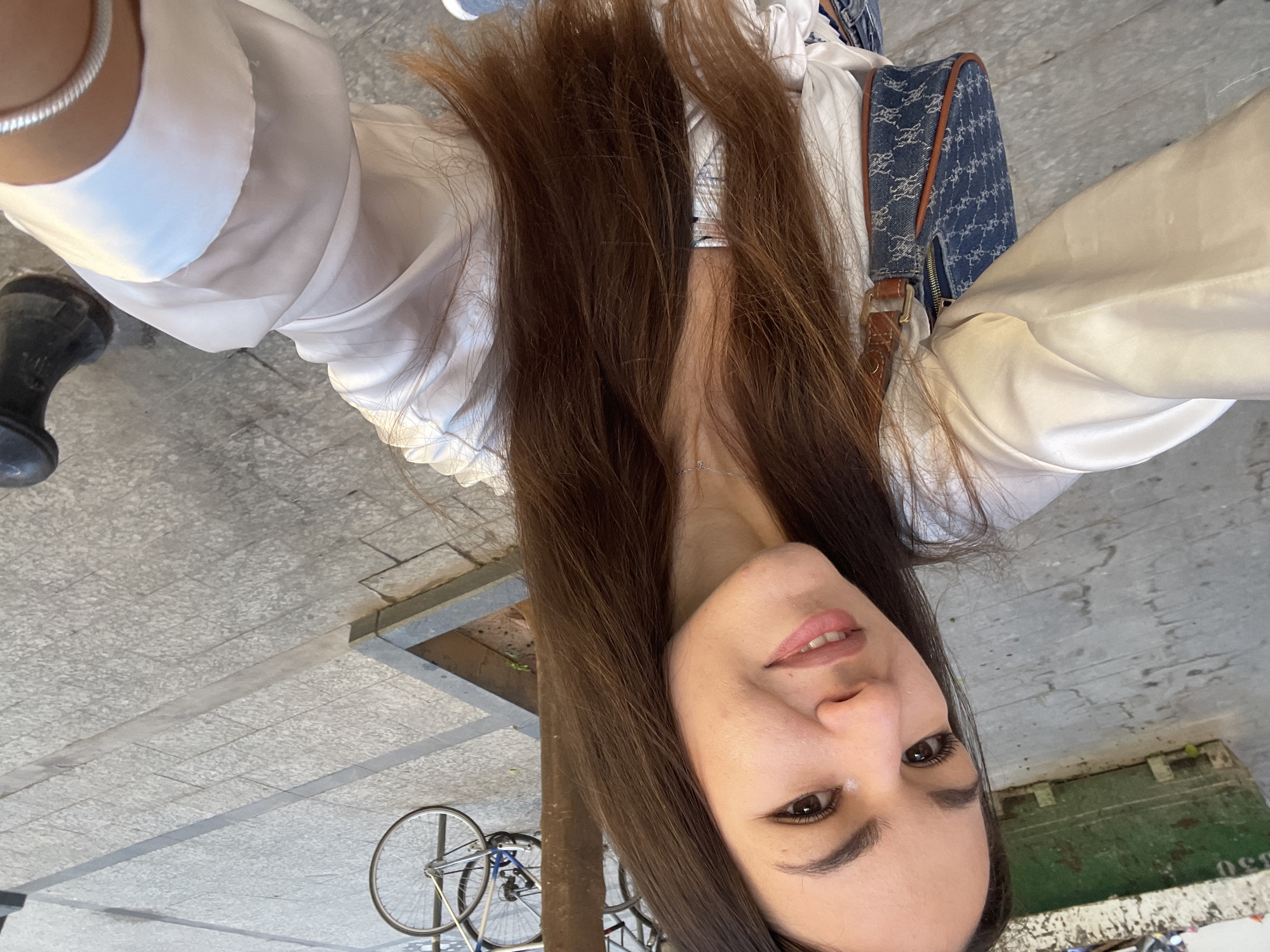 A woman is taking a selfie. She has long brown hair and is wearing a white blouse. A blue patterned bag is slung over her shoulder.
In the background, a bicycle is visible near what appears to be a tree well or planter along a sidewalk. The surrounding area suggests an urban setting.
The location is València, Spain.