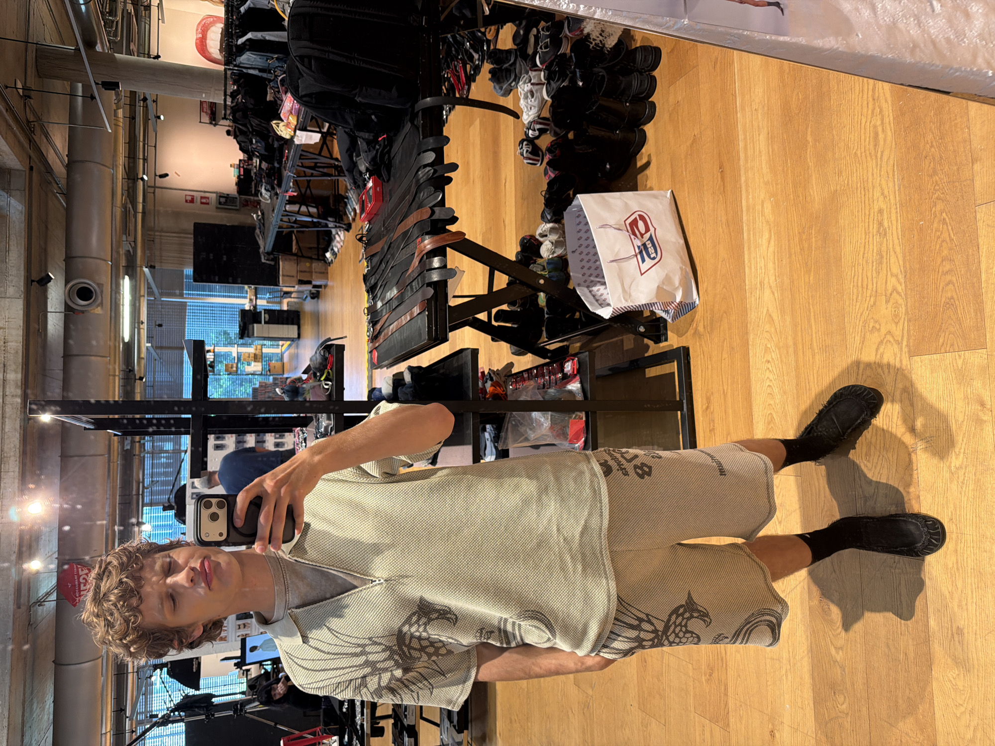 A young man is captured in a mirror selfie within what appears to be a retail store or showroom in Mirabella, Italy. He stands facing the mirror, holding a smartphone to take the picture, with his reflection visible from the waist up. He has curly brown hair and is squinting with a slight pout on his lips, possibly due to the bright lighting or a deliberate expression.
He is wearing a two-piece outfit consisting of a short-sleeved, collared shirt with a half-zip closure and matching shorts. The fabric is light-colored, possibly beige or light grey, with a textured, woven pattern. Dark grey or black stylized eagle or dragon designs are prominent on the front of his shirt and on the sides of his shorts, accompanied by other abstract patterns and faint text elements that are not clearly legible. He wears black socks and black moccasin-style shoes.
The setting features a warm-toned wooden floor. The background reflects an industrial-style interior with high ceilings, exposed metallic pipes, and various light fixtures. To the right, there are racks displaying merchandise, including belts and shelves packed with shoes. A white shopping bag with a red and blue "CPJ" or similar logo is seen on the floor near the merchandise. In the far background, a large artistic image of red lips is visible on a wall. The overall ambiance suggests a modern, urban retail environment.