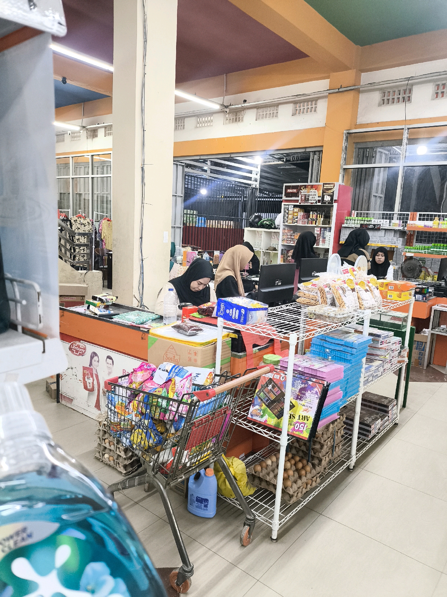The image depicts the interior of a small retail store, likely a minimarket or convenience store, in the city of Posso, Indonesia. The scene is bustling with activity, suggesting it's a busy shopping environment.
At the forefront, a shopping cart is filled with various packaged goods, including snacks and other grocery items. Directly behind the cart, a display rack is laden with more products, such as boxes of crackers, stationery items, and what appear to be school supplies like coloring pencils. Below this rack, stacks of egg cartons filled with eggs are visible.
In the middle ground, there are several individuals, mostly women wearing hijabs, operating at a checkout counter. They appear to be cashiers, attending to customers or processing transactions. One cashier is looking down at a computer monitor, while others are engaged in their tasks. The counter area is organized with products, bags, and payment terminals. A prominent display featuring "Lifebuoy" soap with images of a man and woman suggests personal care products are also sold.
The environment of the store is characterized by bright fluorescent lighting, illuminating the shelves and products. The ceiling has multiple colored panels, with orange and brown being the most prominent. The walls are painted in light colors, and there are visible structural pillars. Towards the back of the store, more shelves stocked with various items are present, and a glimpse of an outdoor area or a service entrance is visible through an open doorway. The overall impression is that of a typical local Indonesian retail establishment.