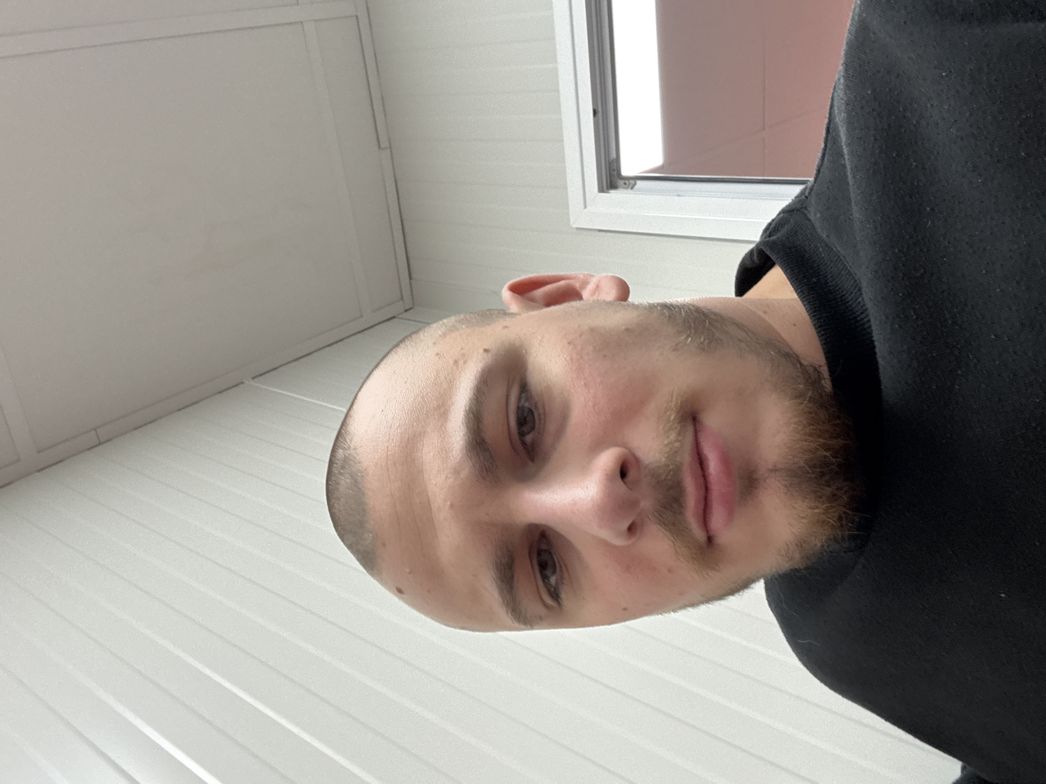 This media file presents a close-up portrait of a young adult male, captured with an unusual 90-degree counter-clockwise rotation. The man occupies the right half of the currently rotated frame, looking directly towards the viewer with a neutral, calm expression. He has a very short buzz cut or shaved head, along with light stubble forming a short beard and mustache. His eyebrows are dark, and his eyes are brown. He is wearing a dark, likely black, long-sleeved top, possibly a sweatshirt or hoodie.
The background consists of light-colored, likely white, walls featuring prominent horizontal paneling or grooves. In what would be the upper right corner if the image were correctly oriented, a window with a white frame is visible, letting in bright, natural daylight. The view through the window appears to be a light exterior, possibly sky or a plain wall. The lighting is even, suggesting daytime. There is no visible text. The scene is an indoor setting located in Stara Zagora, Bulgaria.