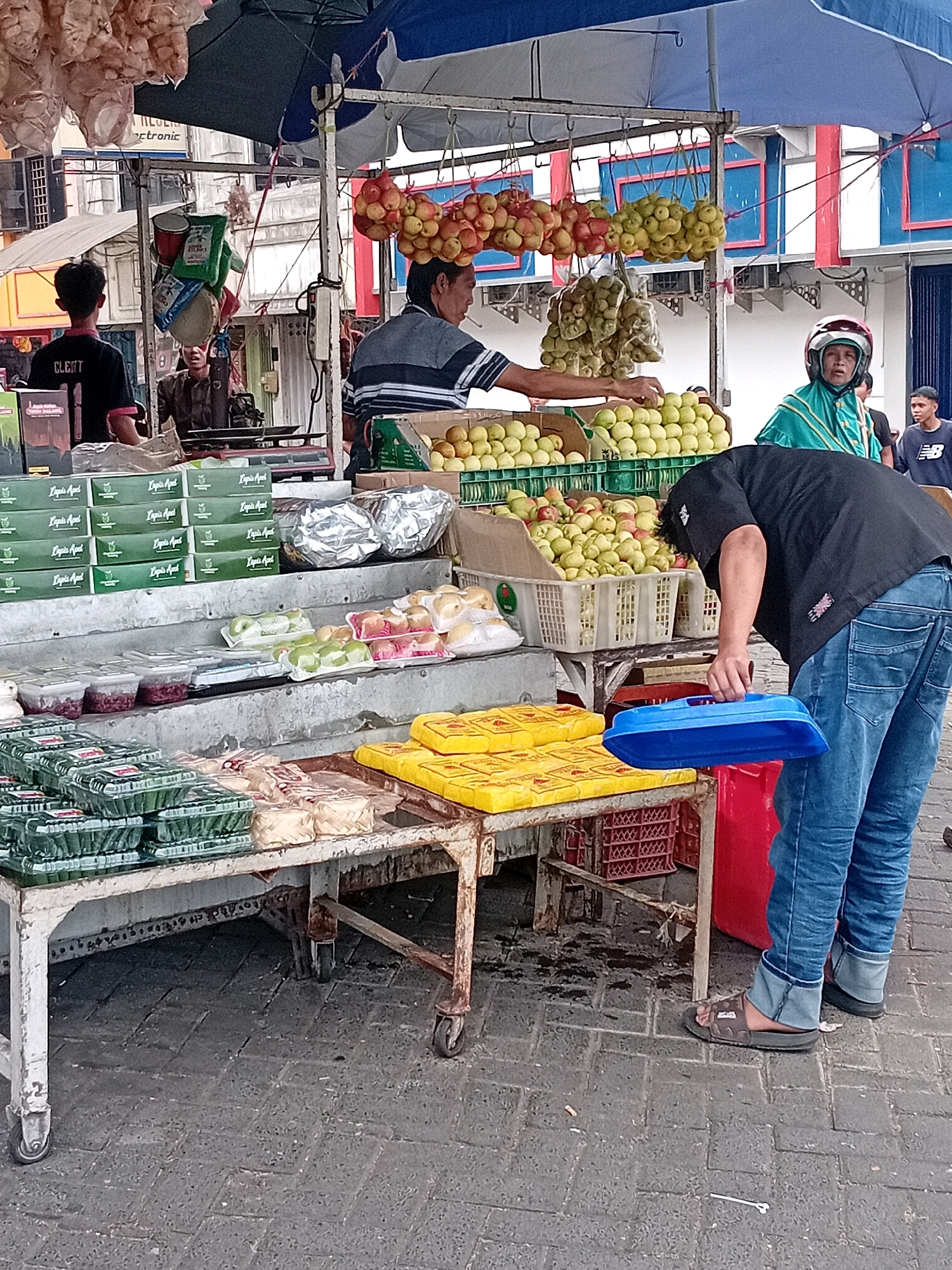 The image depicts an outdoor market scene in Lawang, Indonesia. In the foreground, a person wearing blue jeans and dark sandals is bending down, holding a blue plastic container. To the left of this individual, a white metal table with wheels is laden with various food items packaged in clear plastic containers and woven baskets. Some of these items appear to be green vegetables or fruits. Behind this table, another white metal table is visible, also holding containers of food, including what look like sweets in plastic wrappers.
Further back, a fruit stall is prominent, with bunches of red and yellow apples hanging from the stall's canopy. Baskets of green apples are also displayed. Several people are present at the stall. A man in a striped shirt is interacting with the fruit display. To the right, a person wearing a green hijab and a turquoise outfit is standing near a motorcycle helmet. Another young person in a black t-shirt with a white logo is visible in the background, to the far right. The market appears to be set up on a paved area with a patterned stone surface. Various awnings and building structures are visible in the background, suggesting an urban environment.