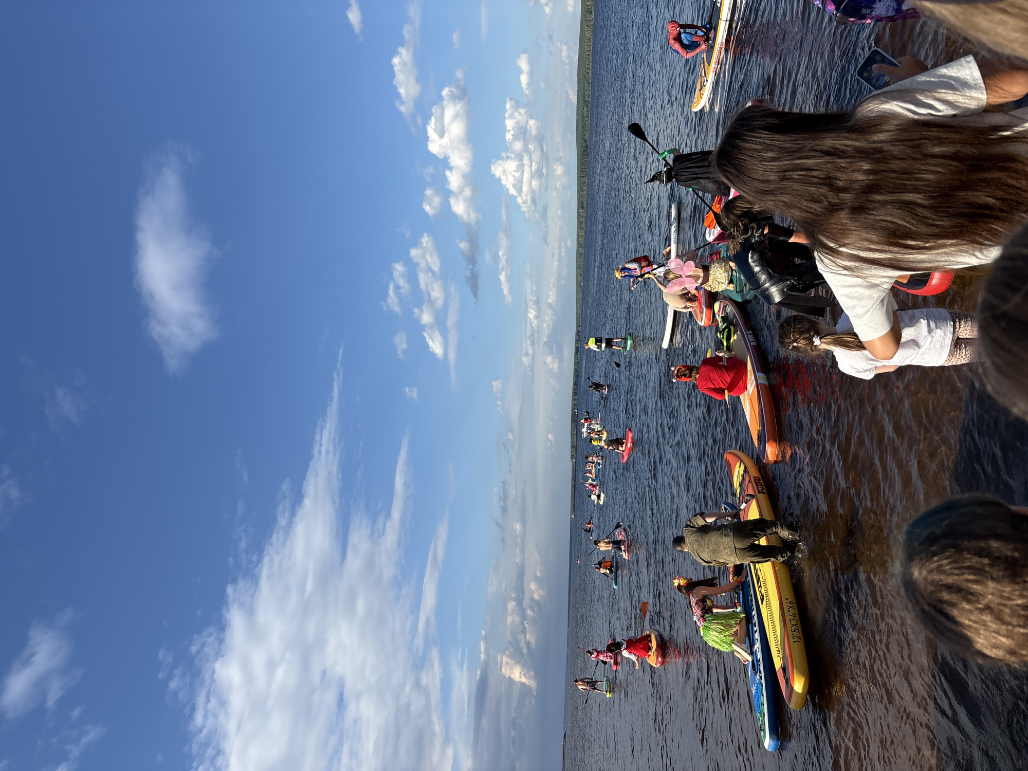 A large group of people are paddleboarding on a body of water, with a forest-lined shore in the background under a blue sky with scattered clouds. Many individuals are dressed in costumes, including what appear to be a Spider-Man costume, a witch costume, and other colorful outfits. The paddleboards are of various colors and designs. Some people are actively paddling, while others are gathered together. The water appears calm. The scene is likely taking place on the Kama River in Chaikovsky, Russia, given the presence of paddleboarders and the characteristic shoreline.