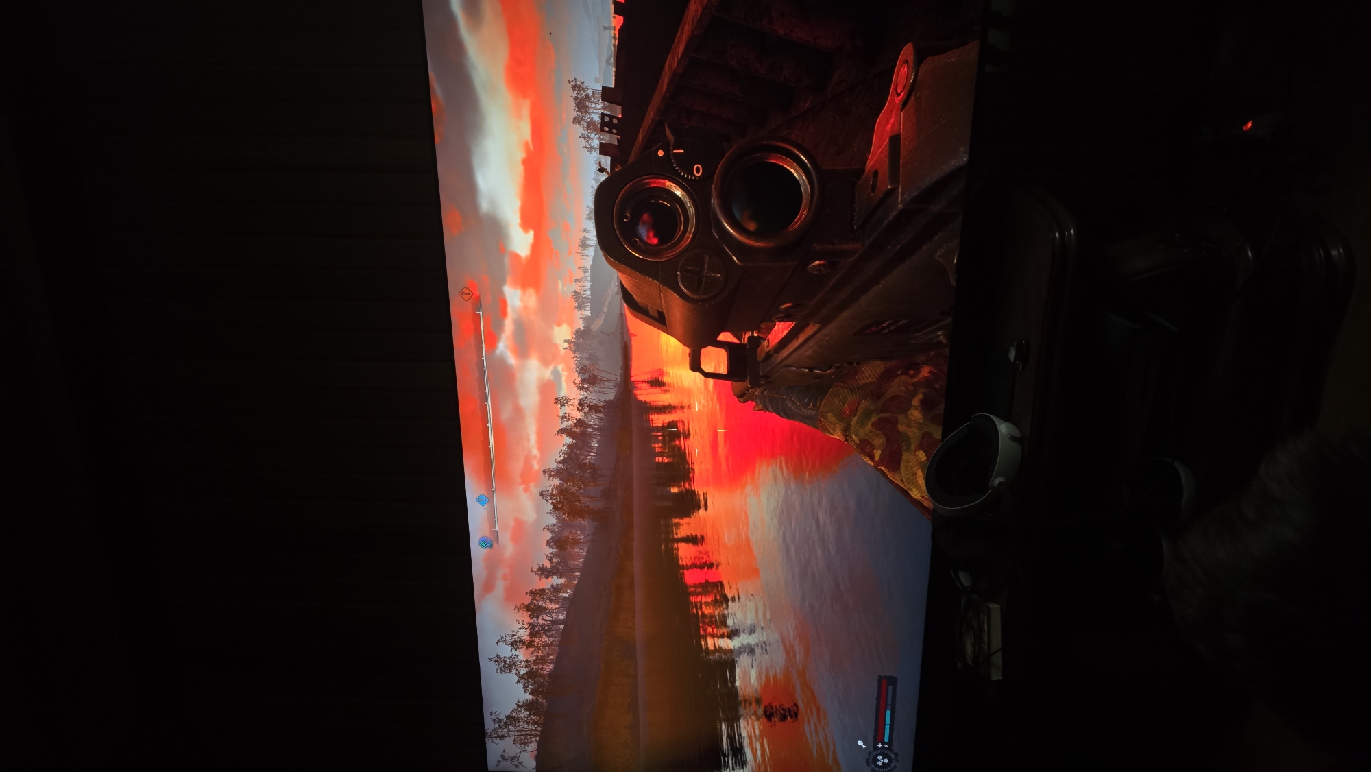 The image is a photograph taken from the perspective of someone playing a video game on a monitor. The game depicts a first-person view of a weapon with a scope, aimed at a serene landscape. The scene is set at sunset, with the sky ablaze in vibrant shades of orange and red, reflecting dramatically on a calm body of water. Silhouetted trees line the distant shore, creating a picturesque and somewhat somber atmosphere.
The video game interface is partially visible on the screen. On the left side, there's a vertical bar, possibly indicating health or a similar game statistic, with small blue icons. At the bottom right, another interface element shows a crosshair and some symbols, typical of a first-person shooter game. The weapon itself appears to be a rifle, with a detailed scope showing a reticle and a numerical indicator, possibly for zoom level. The player's hand, holding the weapon, is clad in a camouflage glove.
The photograph captures the monitor screen, and the surrounding environment is dark, suggesting the room is dimly lit to enhance the viewing experience. Two VR headsets are visible in the lower right portion of the image, resting on a dark surface, hinting that the monitor might be part of a gaming setup. The overall impression is one of a peaceful yet potentially intense gaming session, juxtaposing the beauty of the virtual sunset with the implied action of the game. There are no discernible people within the game's scene, and the visible text is limited to game interface elements. The image is oriented vertically, which may be due to how the photograph was taken or how the game is being displayed.