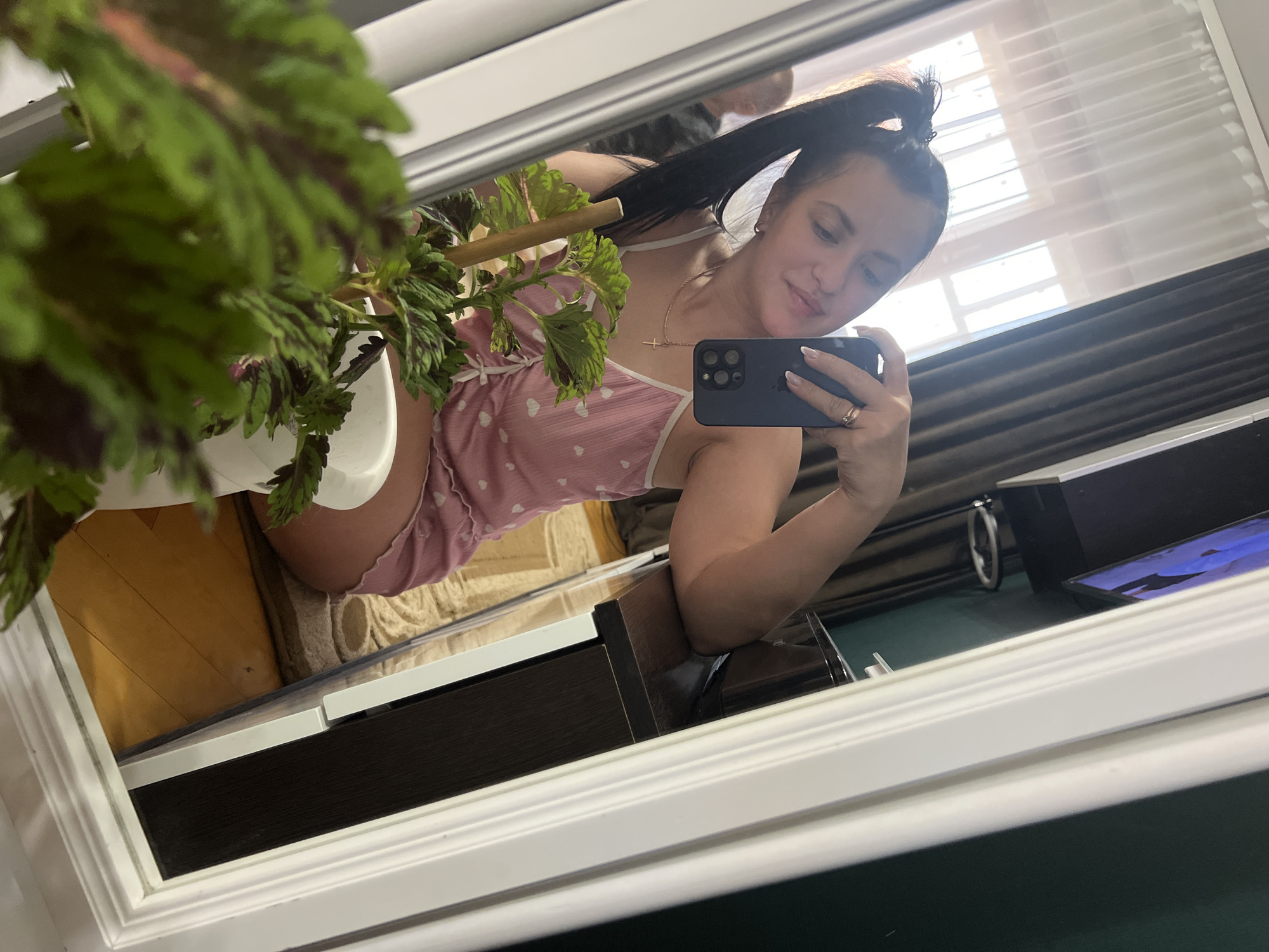 A woman is taking a selfie in a mirror. She has her hair in a ponytail and is wearing a pink slip with white hearts. She is also wearing a necklace with a cross pendant and a ring on her left hand. In front of her, there is a plant in a white pot. The background shows a room with a window, curtains, and a dresser. The floor is made of wood. The woman is sitting down, and her legs are visible.