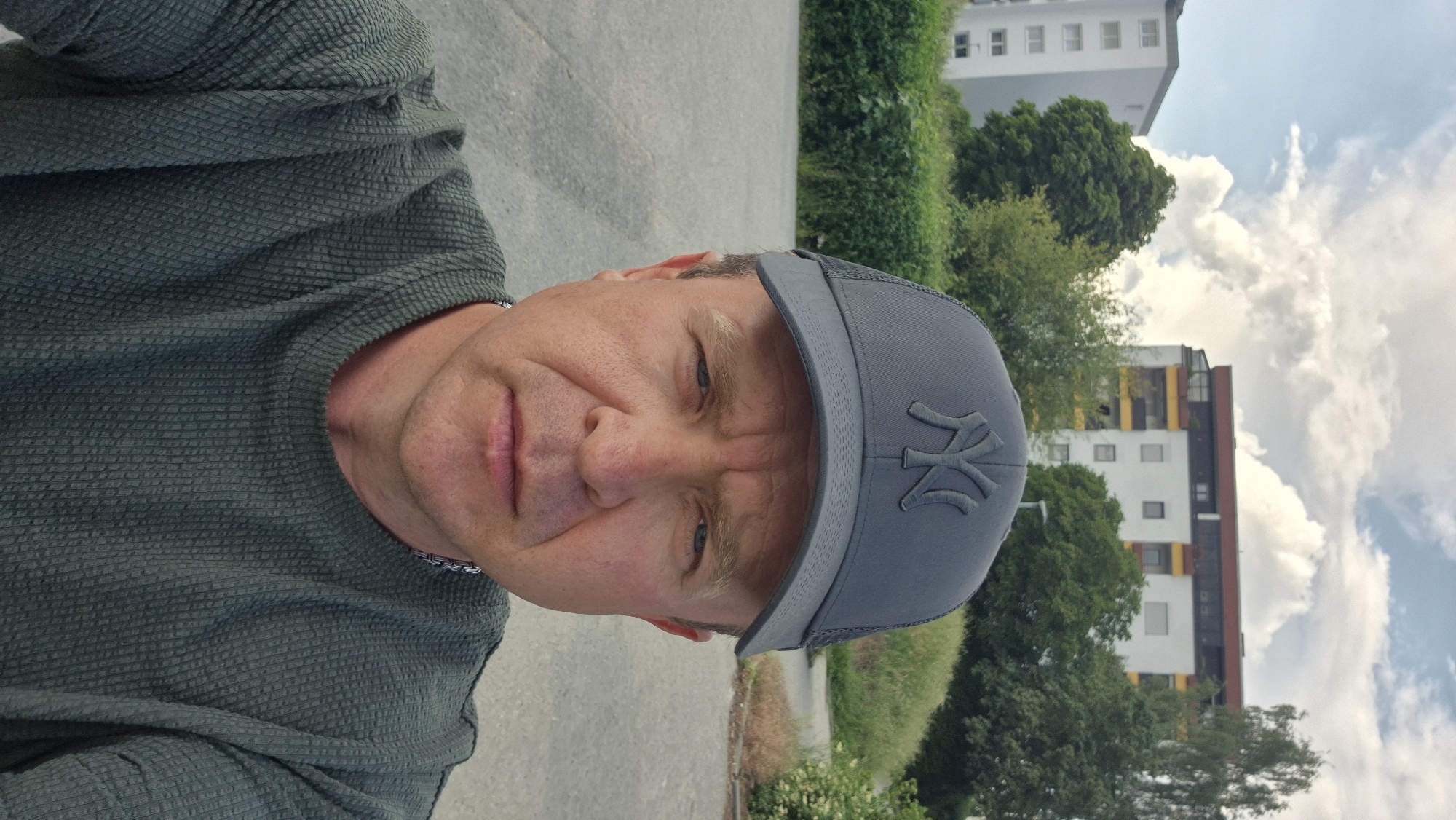 A middle-aged man wearing a dark green textured shirt and a grey New York Yankees baseball cap is looking directly at the camera. The photo is taken from a low angle, suggesting the photographer is at a lower level. The background includes an asphalt road and greenery, with apartment buildings and a partly cloudy sky visible in the distance. The location is likely in Ergolding, Germany, based on the apartment building styles and the urban setting.