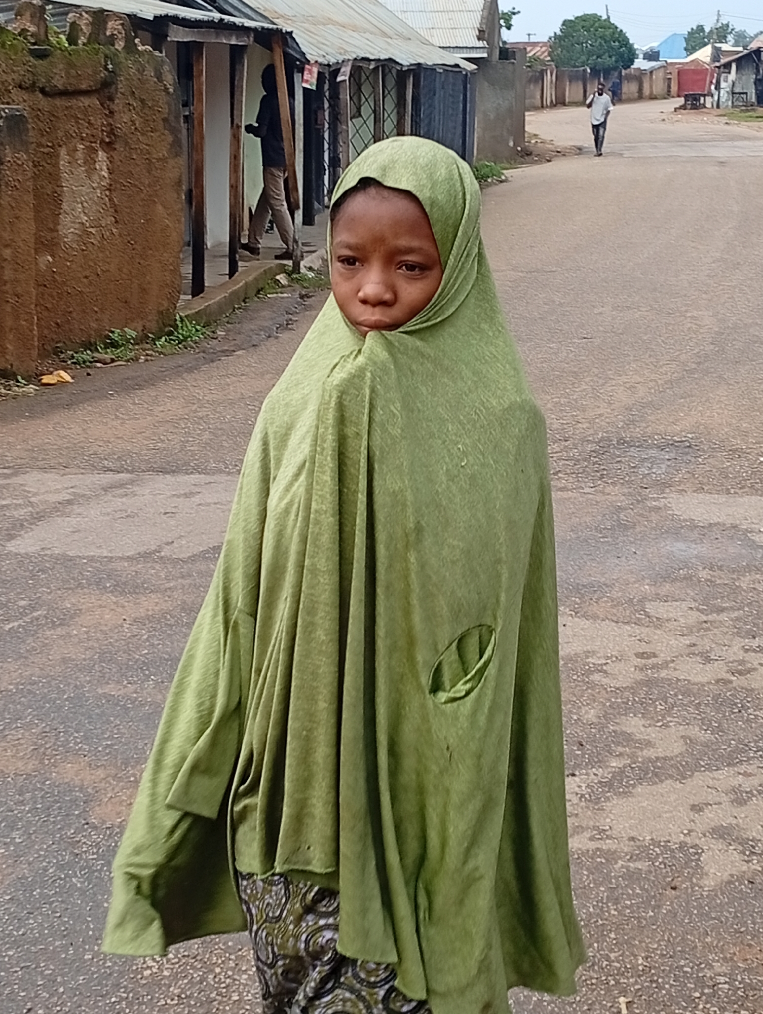 A young girl wearing a green headscarf stands on a dirt road in Jos, Nigeria. The girl's face is visible, and she appears to be looking down or to the side. She is wearing a patterned skirt underneath the headscarf, which drapes over her body. To the left of the girl, there is a rough, textured wall and what appears to be a building with a corrugated metal roof. In the background, a man walks away from the camera on the dirt road. Further in the distance, there are more buildings and trees. The overall impression is of a street scene in a rural or semi-urban area.