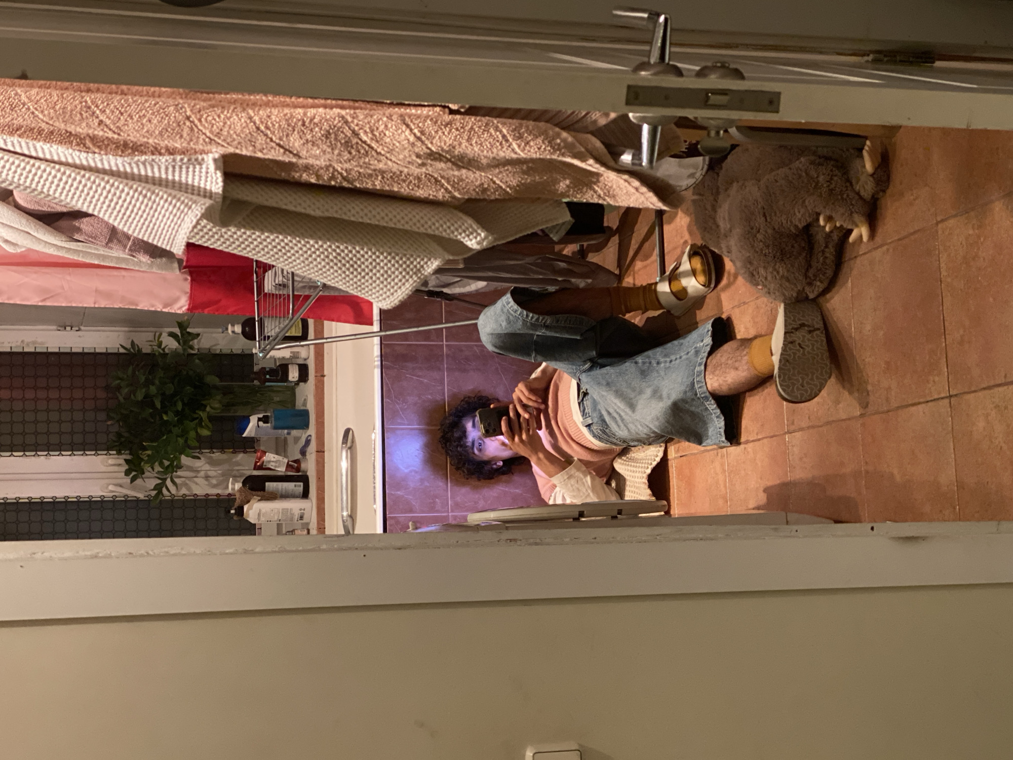 A person is lying on the floor, illuminated by a blue glow from a mobile phone screen. The individual has curly dark hair, wears a pink top, and has light blue jeans and sandals. A pile of towels and other items sit on a clothes airer next to them. There is brown flooring in the room. The scene is viewed from inside a doorway. The location is not discernible from the image.