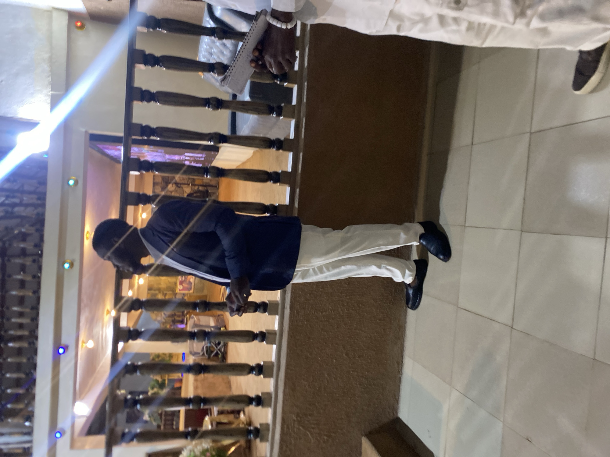 A man stands on a tiled surface, near a brown-colored railing with wooden spindles, in what appears to be a building along Rukuba Road, Nigeria. He is wearing a dark blue blazer and white pants with black shoes. He stands next to a person wearing a white top and holding a small notebook. The background shows a building interior with lights and decorations.