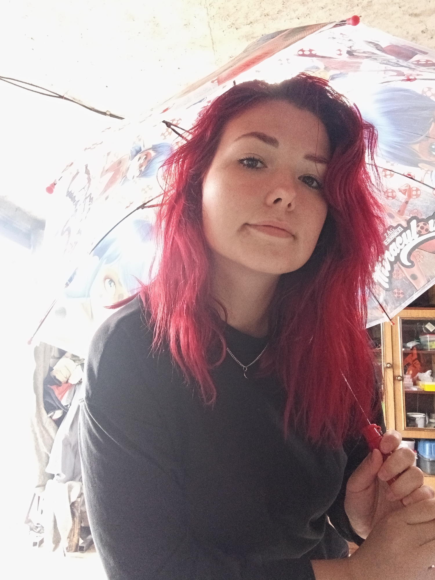 A young woman with vibrant red hair and fair skin is positioned centrally, looking directly at the camera. She is wearing a black, long-sleeved shirt and a silver necklace with a crescent moon pendant. She is holding a partially opened, transparent umbrella with a cartoon print, possibly featuring characters from "Miraculous Ladybug." The umbrella is held above her head and obscures the upper part of the scene.
The background is brightly lit, suggesting a well-lit interior or an outdoor location with strong light. To the right, a portion of a wooden cabinet with glass doors is visible, containing various items on shelves. To the left, there appears to be a cluttered area with clothing or fabric. The wall behind her is textured and light-colored.
The overall lighting is bright, with some areas overexposed, particularly in the upper left quadrant. The image has a casual, selfie-like quality. There are no visible clues indicating a specific location in the city of Chaikovsky, Russia, or any other identifiable geographical context.