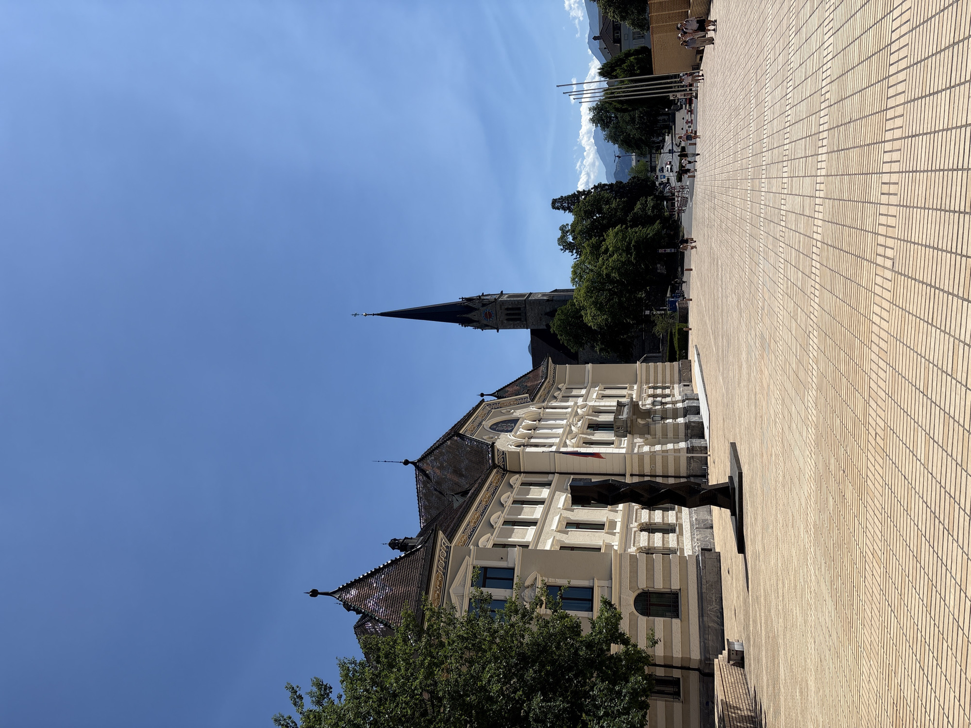 In Vaduz, Liechtenstein, a broad, paved plaza faces the iconic Saint Florin Cathedral, its tall, dark steeple dominating the sky. To the left, the ornate facade of a cream-colored building, likely a government or administrative office, features a steeply pitched, dark tiled roof with decorative accents and dormer windows. A modern, dark spiral sculpture stands in front of this building, casting a shadow on the light-colored, patterned paving stones of the plaza. A small Liechtenstein flag is visible on the building. Beyond the cathedral, a tree-lined street recedes into the distance, with cars and pedestrians present. The background reveals rolling hills and mountains under a clear blue sky. Several people are walking on the plaza and the street, appearing to be tourists or locals enjoying the sunny weather.
