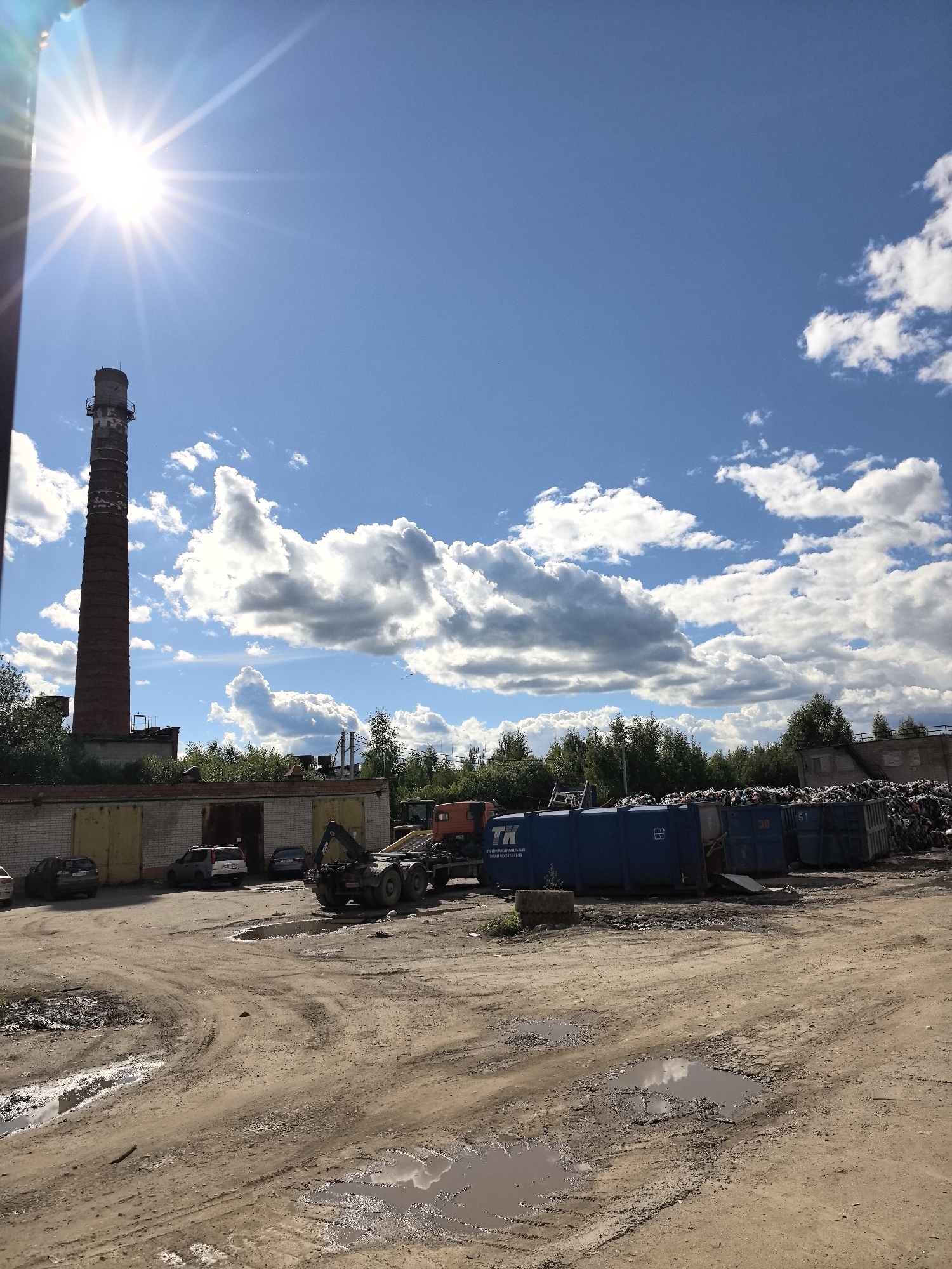 The image depicts an outdoor industrial or waste management area under a bright blue sky with scattered white clouds. A tall, reddish-brown brick smokestack with a metal platform near its top stands prominently on the left side of the frame. In front of the smokestack, a low-rise building with a light-colored brick facade and multiple garage-like doors is visible, with several parked cars in front of it.
The central and right portions of the image show a collection of large, blue dumpsters, some labeled with numbers like "30" and "51," filled with various types of waste. A truck with a crane attachment is positioned behind some of these dumpsters. Beyond the dumpsters and the truck, a dense line of green trees forms the horizon, with additional industrial-looking structures visible through the foliage on the right. The ground in the foreground is unpaved, with areas of dirt, mud, and visible tire tracks, indicating a working environment. The sun is shining intensely, casting a bright glare and lens flare in the upper left quadrant of the image. The scene appears to be located in a rural or industrial outskirts area, consistent with a waste processing or recycling facility.