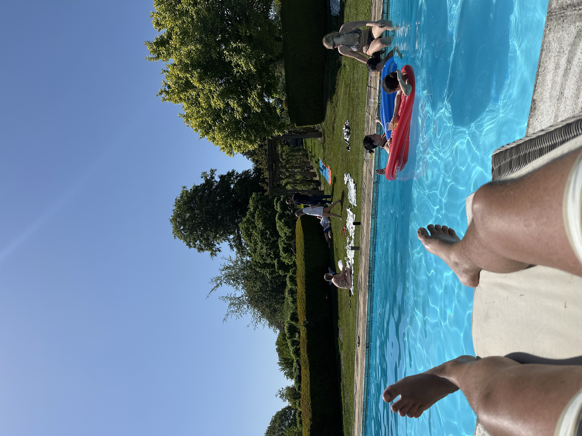 The image depicts a scene at a swimming pool in a backyard setting, likely in a residential area. The sky is clear and blue, suggesting a sunny day. The pool is the main subject, with multiple people either in the pool or standing near it. Some people are using inflatable floats. Green grass surrounds the pool area, and a trimmed hedge separates the pool area from the rest of the backyard. There is a pergola structure visible in the background. The foreground shows a person sitting beside the pool, with their feet dangling over the edge. There are no specific details suggesting a particular city location.