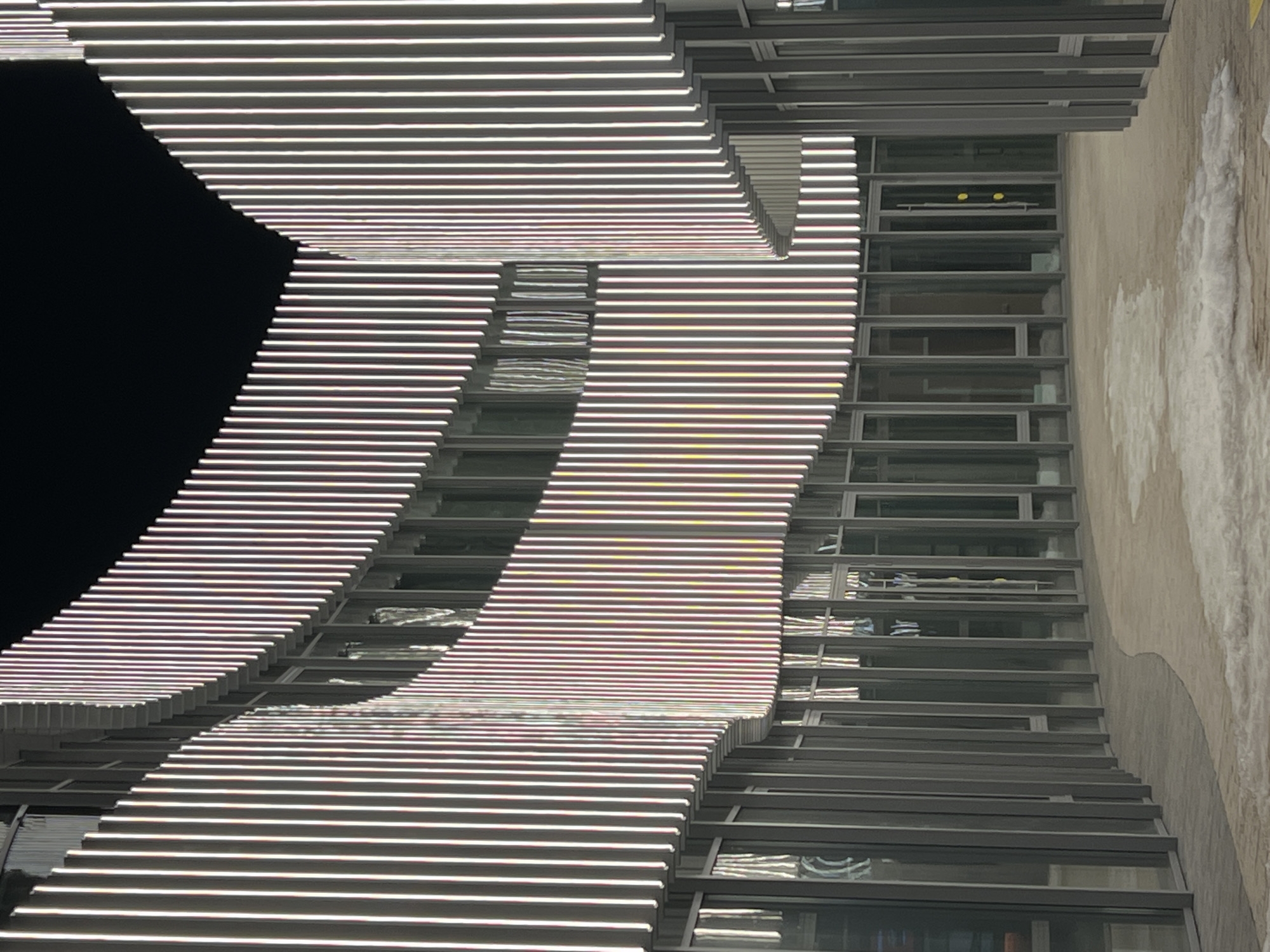 The image captures an exterior nighttime view of the IT Park named after Bashir Rameev in Kazan, Russia. The scene depicts a modern architectural complex with a prominent curved facade. The upper sections of the building feature numerous vertical, evenly spaced, illuminated light strips or fins. These elements predominantly emit white light, exhibiting subtle chromatic shifts to pink, violet, and yellow hues across their surfaces. Below these illuminated elements, the building's lower levels are clad in a continuous series of floor-to-ceiling glass panels, framed by slender dark grey vertical mullions and horizontal transoms, indicative of commercial or office spaces. A smooth, light-colored paved pathway or courtyard curves in the foreground, mirroring the building's contour. Irregular patches of partially melted snow are scattered across this pathway. The sky above the building is uniformly dark. No individuals or discernible activities are present in the scene.