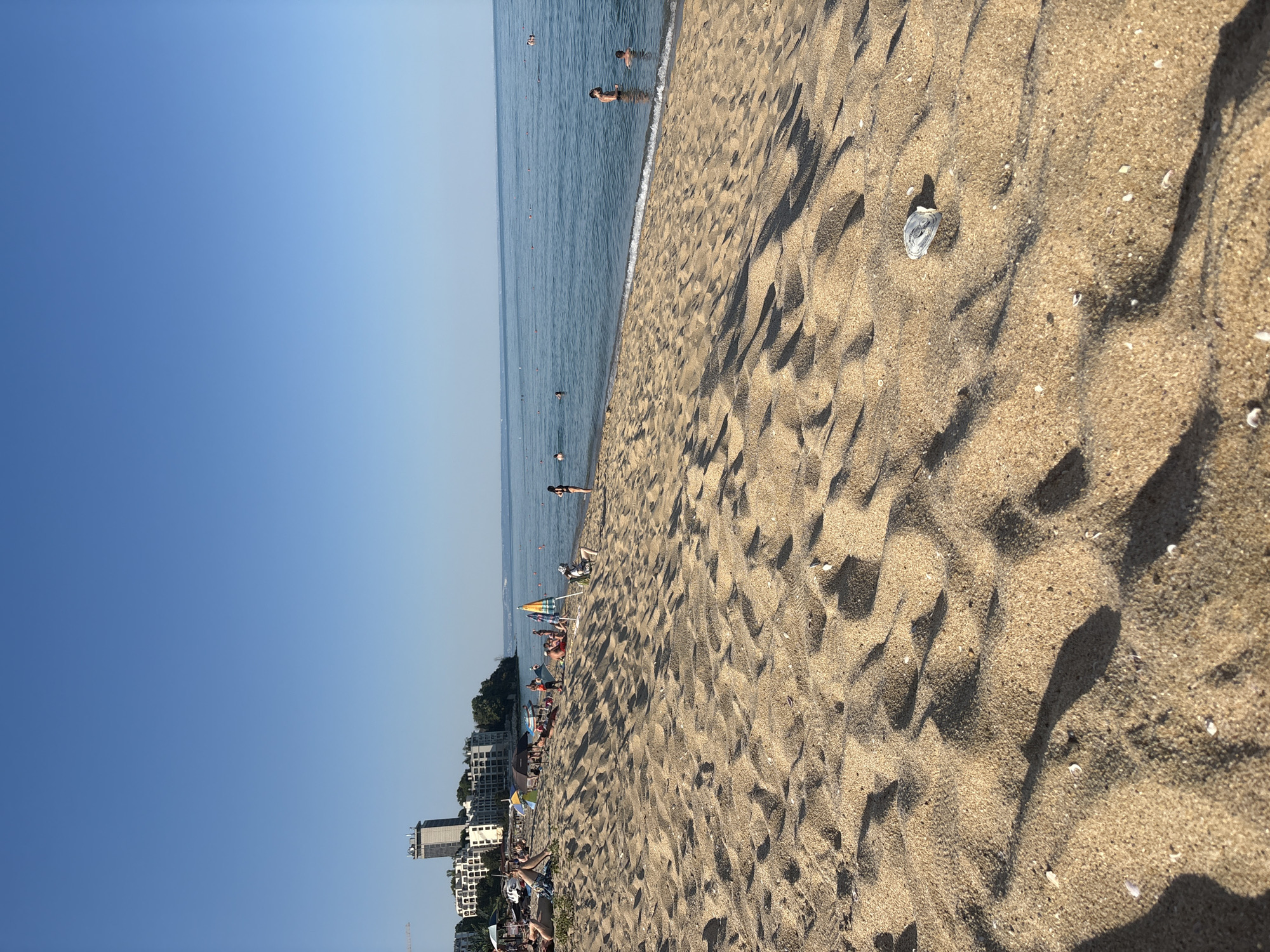 The image depicts a crowded beach scene under a clear blue sky, suggesting it is daytime and the weather is pleasant. The perspective is from the sandy beach, looking towards the sea and a line of buildings along the coast.
In the foreground, the sandy beach is textured with ripples and shadows, with a few small shells scattered across its surface. A discarded plastic bottle is visible on the sand.
Further back, the sea is calm and blue, with several people visible swimming or wading in the water. Closer to the shore, the gentle waves create a white foam line.
Along the coastline, a strip of beach is occupied by numerous people relaxing under umbrellas, sitting on chairs, or walking. Colorful beach umbrellas are dotted across the sand, adding vibrancy to the scene. Beyond the beachgoers, a cluster of buildings, appearing to be hotels or apartments, lines the coast. Some of these buildings are tall, modern structures. Lush green vegetation is interspersed with the buildings.
The overall impression is of a busy summer day at a popular seaside resort, with people enjoying leisure activities. The image captures a bright, sunny atmosphere, characteristic of a holiday destination. No visible text is present in the image.
