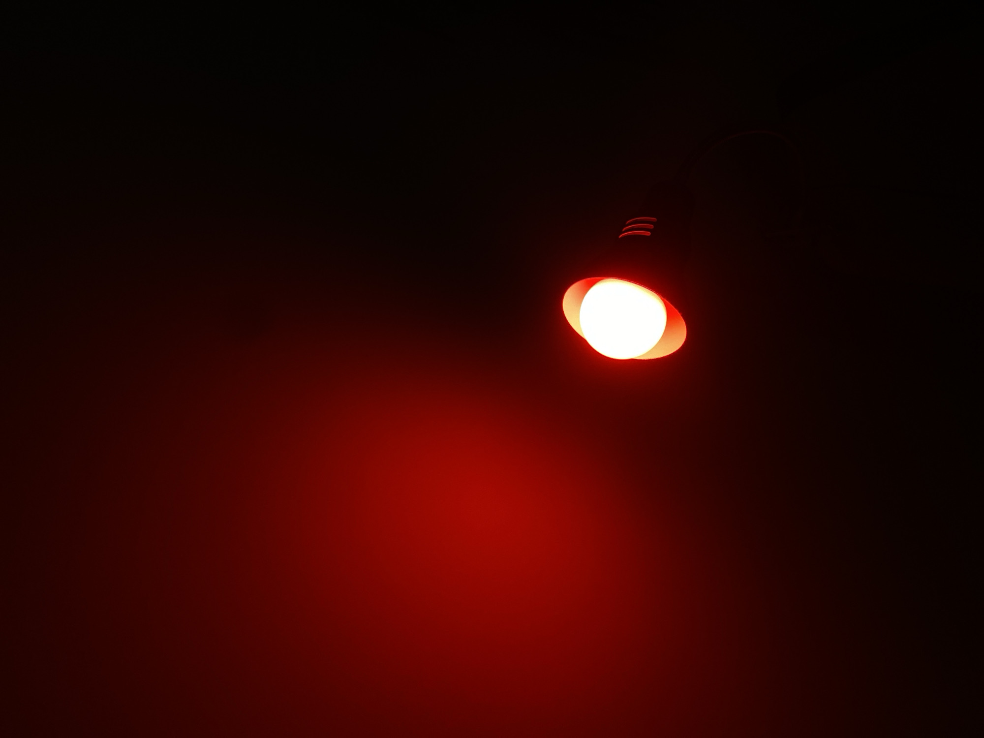 The image captures a single, bright red light source against a dark, ambient background. The light is circular with a warm, glowing effect, suggesting it might be an illuminated object like a signal or a bulb. To the right of the light, there are faint, curved red streaks, possibly an artifact of the lighting or a secondary light source. The overall scene is dominated by shades of deep red and black, creating a dramatic and focused effect on the central light. There are no people or specific city landmarks visible in the image, and therefore no identifiable location within Budva, Montenegro.
